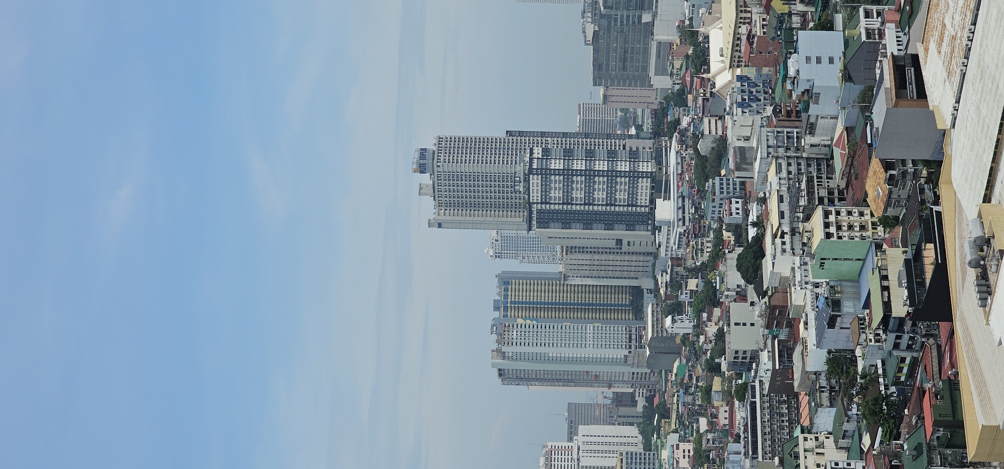 An aerial, medium shot captures a sprawling cityscape under a bright, pale blue sky with wispy clouds. The scene is dominated by a dense collection of buildings, ranging from mid-rise structures to towering skyscrapers, all tightly packed together. The architecture is diverse, featuring modern glass and steel high-rises alongside older, more utilitarian buildings with visible concrete and metal roofing.
Several prominent skyscrapers stand out, with one in the center-right displaying the text "ELITE TOP" on its upper facade. The lower portions of the image reveal a denser urban fabric, characterized by a mix of residential and commercial buildings with varied roof colors, including red, green, and gray. Some buildings appear to be under construction or in disrepair, indicated by exposed structural elements. The overall impression is of a densely populated and developed urban environment, likely in the business district of Makati, Philippines, given the architectural styles and density typical of such areas. No people are visible in the frame.