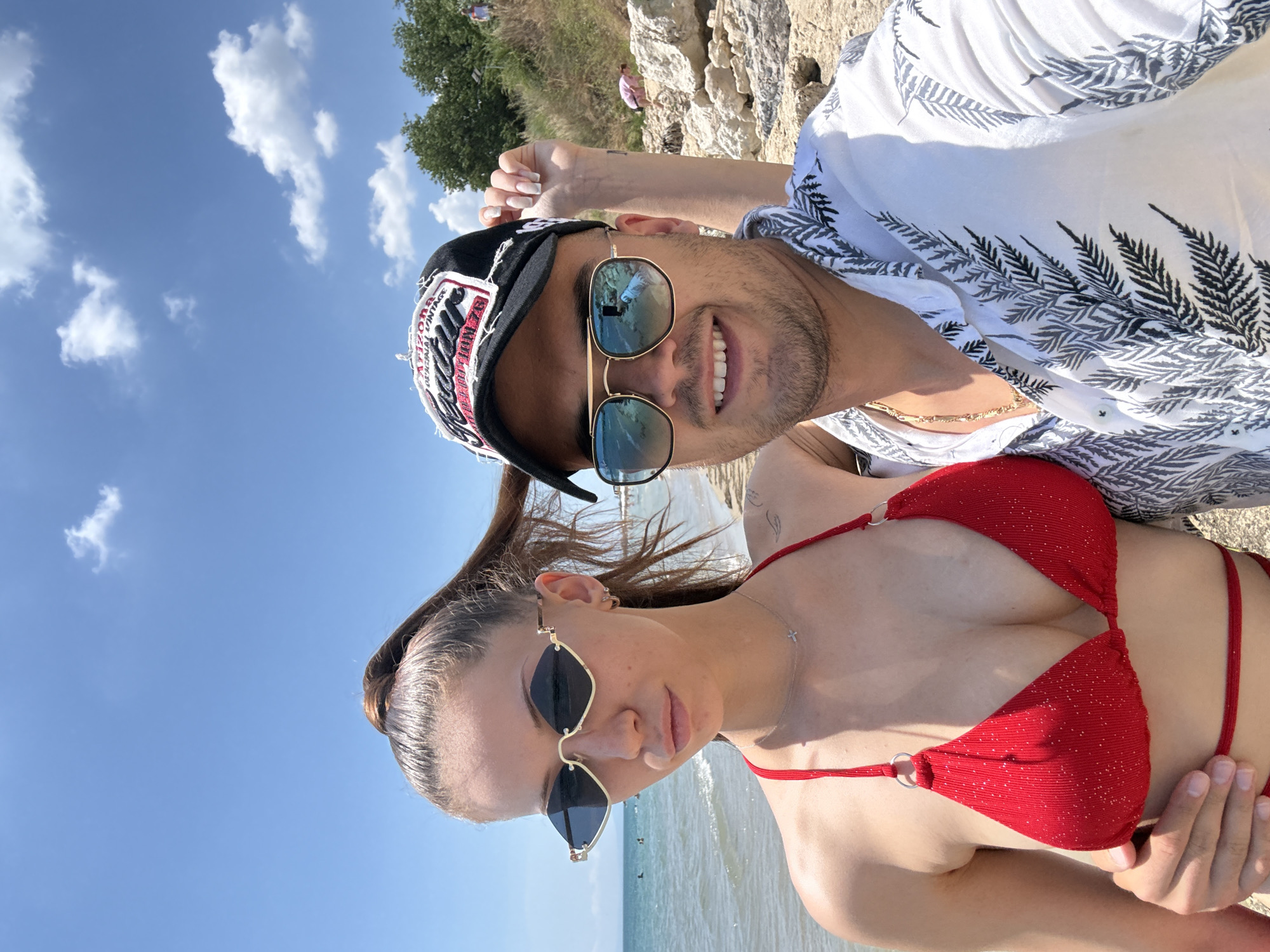 This is a vertically oriented selfie of a man and a woman on a beach. The man, in the foreground on the left, is wearing a black baseball cap with red and white lettering, sunglasses with gold frames, and a white shirt with a black leaf pattern. He is smiling and looking at the camera. The woman is on the right, wearing a red bikini top and sunglasses with dark lenses and gold frames. Her hair is pulled back in a ponytail.
The background shows a clear blue sky with scattered white clouds, and a body of water, likely the sea, with a few small birds on it. Behind them, to the left, is a grassy hill with trees, and to the right are rocky formations. The lighting suggests it is daytime, likely sunny. The overall mood appears relaxed and happy, typical of a beach outing.
Visible text on the man's cap appears to be "ARIZONA VINTAGE". There is also a small cross pendant necklace visible on the woman.