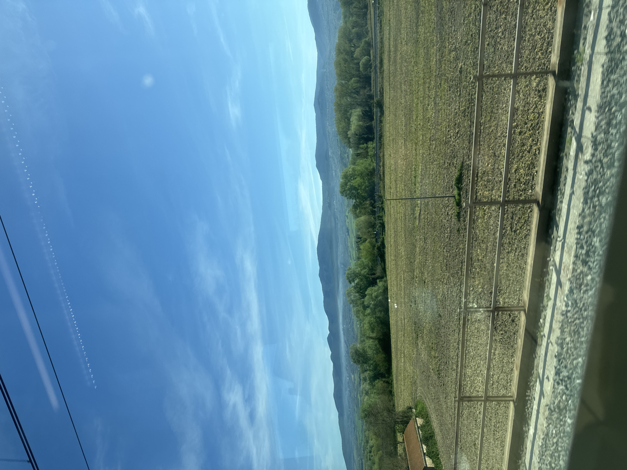 The image depicts a view from a train window in Alviano Scalo, Italy. The sky is blue with streaky clouds and contrails, visible in the upper left. Parallel power lines are visible in the far-left edge of the frame. To the right of the sky is a landscape featuring a line of trees along the base of a mountain range. A field, separated by a wire fence with horizontal rails, stretches across the right side of the image. The field appears freshly tilled. Gravel and possibly a concrete wall are visible in the foreground, immediately adjacent to the train window. The entire scene is viewed at a slight angle.