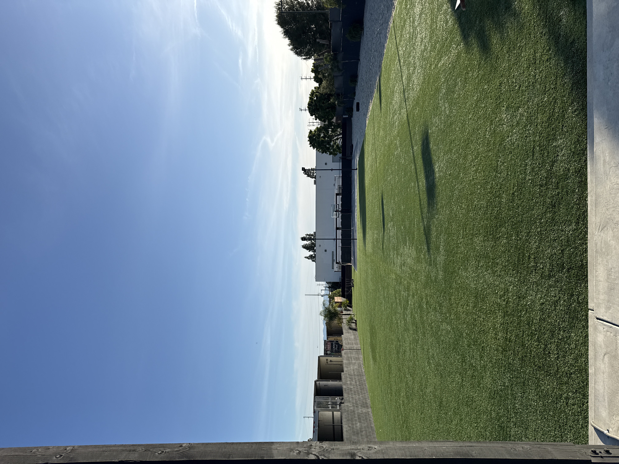 The image shows an outdoor scene with a clear blue sky and some wispy clouds. A patch of artificial green turf is visible on the right side of the frame. Adjacent to the turf is a gray concrete wall or structure. Beyond the turf and wall, there appear to be storage containers or metal structures. In the background, there's a white building with a basketball hoop visible. A row of trees and vegetation lines the area behind the building. Utility poles and wires are also present in the background. The location appears to be in an urban area of Los Angeles, United States of America, given the presence of the concrete structures, utility infrastructure, and the types of buildings visible in the distance. The image has been rotated or taken with a phone rotated sideways, causing the elements to appear on their side.