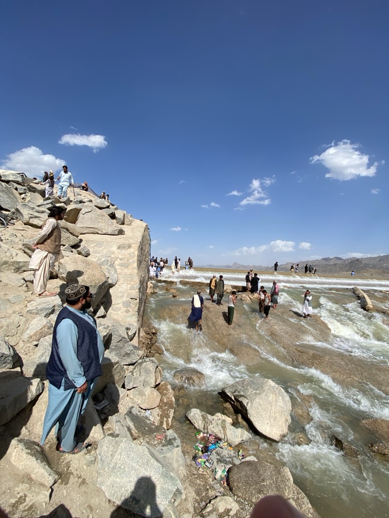 The image captures a scene in what appears to be the city of Kim, Afghanistan, during daylight under a partly cloudy sky. A group of people are gathered on a natural rock formation, with water flowing down the surface. Some individuals are standing on the rocks, while others are walking or wading through the water. The terrain is rocky and rugged. Some of the men in the image are wearing traditional clothing, including vests and headdresses. In the foreground, a man is standing on the rocks, wearing a light blue outfit with a dark vest. There is litter, including what appears to be plastic wrappers and containers, near the water's edge. In the background, hills or mountains are visible on the horizon.