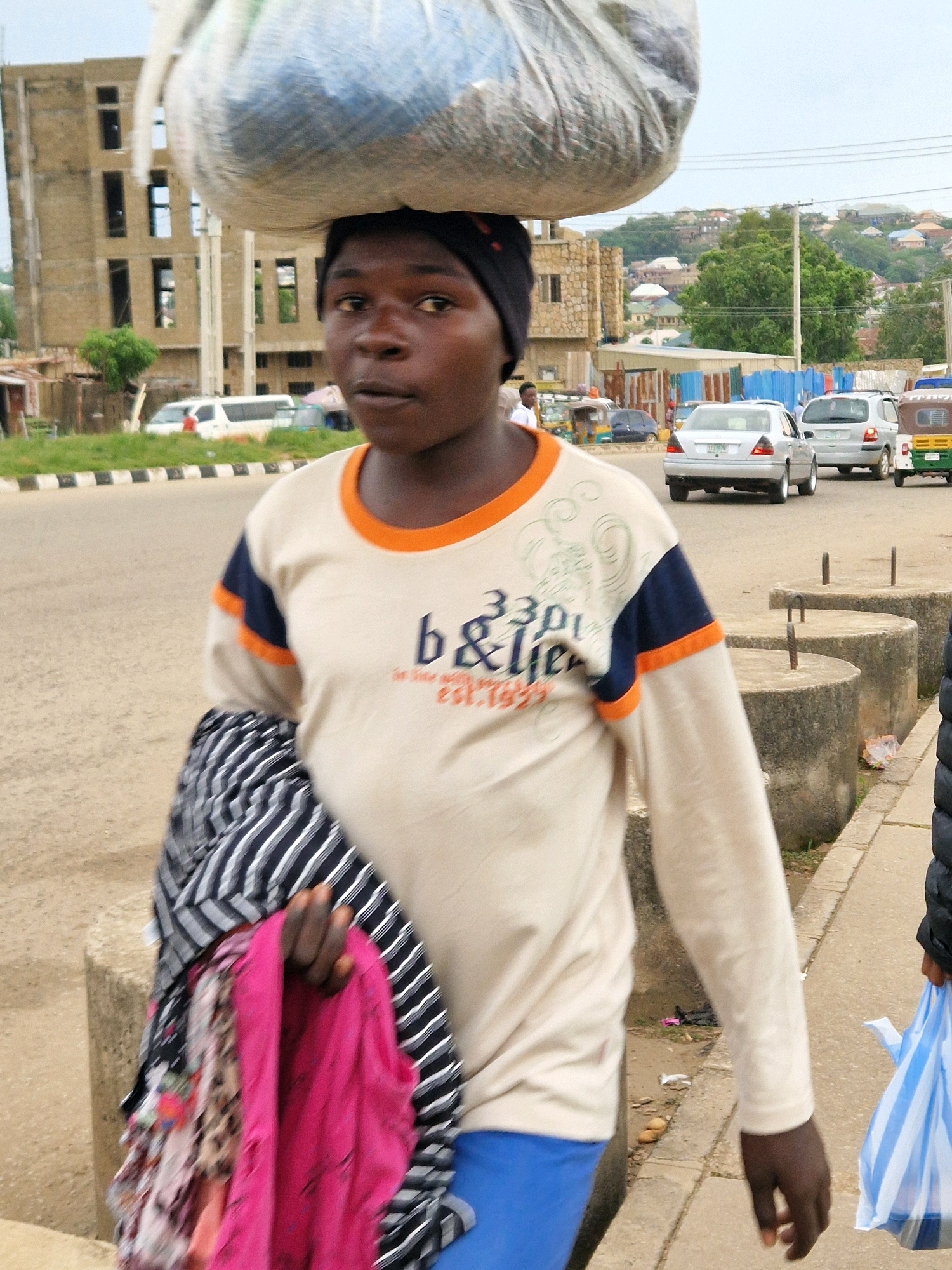 A young person with dark skin walks with a large bundle of items on their head, balanced with their neck. They are wearing a light-colored long-sleeved shirt with orange and navy trim and blue pants. They are also carrying a bundle of clothes in their left arm. The scene appears to be in Jos, Nigeria, with a busy street in the background. The street has several cars, including a silver sedan and a white SUV, and what looks like a rickshaw. A multi-story building under construction is visible on the left side of the image. The ground is paved with asphalt, and there are concrete bollards lining the sidewalk. The sky is overcast.