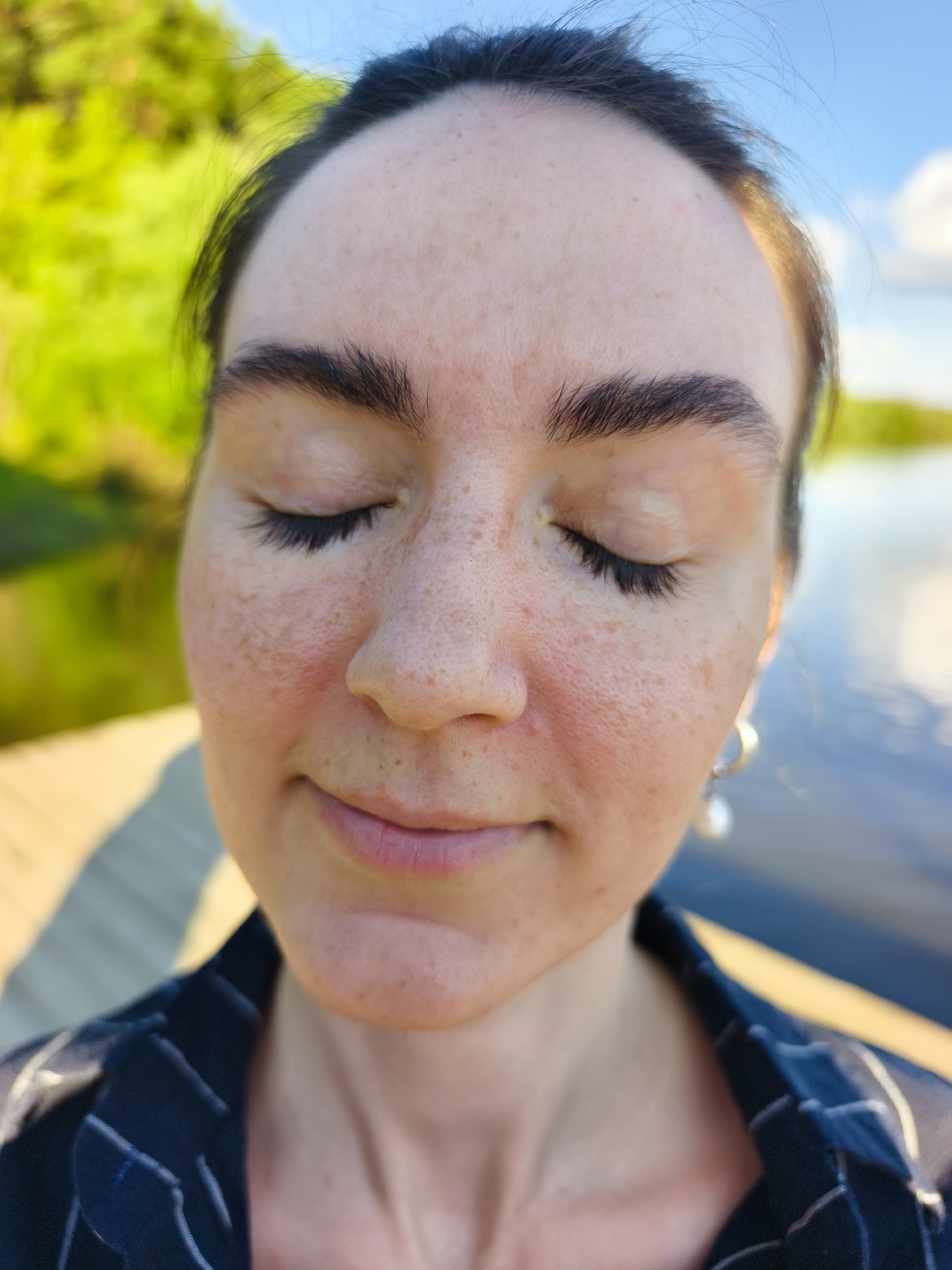 The image is a close-up, eye-level, outdoor portrait of a woman with her eyes closed, facing slightly upwards. She has fair skin with numerous freckles across her forehead, cheeks, and nose. Her eyebrows are dark and well-defined, and her eyelashes are long and dark. She is wearing a dark blue collared shirt with thin white pinstripes and a pearl earring in her visible right ear. The background is blurred, showing a body of water with reflections and a line of trees or greenery to the left. A wooden surface, possibly a dock or deck, is visible in the lower left quadrant. The overall lighting suggests it is daytime. The location is identified as Onochino, Russia.