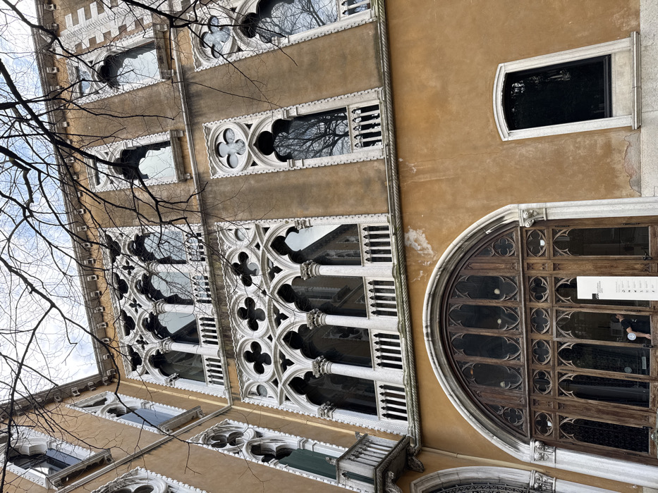 The image displays a portion of the Venetian Gothic facade of Ca' Corner della Regina, located at Santa Croce 2215, Venice, Italy, viewed from a low angle looking upwards. The building features an ochre-colored stucco exterior with significant white Istrian stone decorative elements, including window frames, tracery, columns, and balustrades.
The visible ground floor presents a large, central arched wooden entrance or window system composed of multiple panels with intricate Gothic tracery, including quatrefoil motifs. To the immediate right of this, a rectangular window with a simple white stone surround and sill is present. A tall, rectangular white sign with black text and graphics stands upright on the ground in front of the large wooden entrance. Inside the wooden entrance, a single individual wearing a dark short-sleeved top is partially visible, bent forward, appearing to interact with an unseen object or display.
The first and second floors above exhibit characteristic Venetian Gothic architecture. Each features a prominent triple-arched loggia or window with slender white stone columns supporting pointed arches, surmounted by ornate quatrefoil tracery. A white stone balustrade runs along these first and second-floor features. Additional, narrower windows with pointed arches and quatrefoil details are visible on either side of the main loggias, also with smaller white stone balustrades.
The upper portion of the image shows an overcast sky, partially obscured by numerous bare tree branches. Reflections of these branches and the sky are visible in the building's dark windows. The ochre facade exhibits some areas of weathering, discoloration, and minor flaking paint.