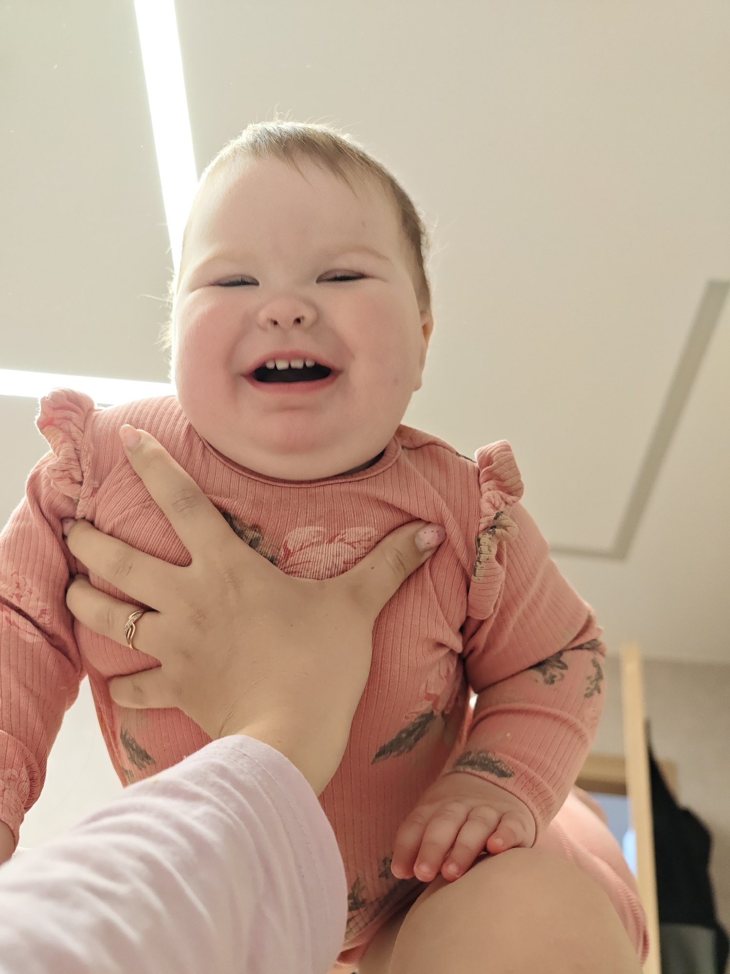 The image presents a close-up, low-angle view of a baby being held by an adult. The main subject is a happy baby, likely an infant or young toddler, with light hair and chubby cheeks, positioned towards the top of the frame. The baby is wearing a light pink, ribbed long-sleeved onesie with ruffled detailing on the shoulders and a subtle pattern of dark feather-like or floral motifs. Its face is beaming with a wide, open-mouthed smile, revealing small teeth, and its eyes are squinted in an expression of joy.
An adult's hand is visible in the foreground, supporting the baby's torso. This hand wears a gold-colored ring with small embedded stones on the fourth finger. The adult's light pink long-sleeved top is also partially visible. The activity captured is an affectionate and playful interaction, with the adult holding the baby up, possibly during playtime.
The setting is an indoor space, likely a brightly lit room in a home. The background is mostly light-colored walls and ceiling, featuring a prominent, bright linear light fixture overhead. In the upper right background, a slight architectural detail resembling a ceiling panel is visible. Further to the right, a wooden frame, possibly a door or mirror, and a dark hanging item suggest a common room environment. The lighting is bright, indicating a well-lit interior, possibly daytime. The dominant emotion conveyed is pure happiness from the baby, creating a joyful and heartwarming scene. There is no visible text in the image. The location context for this scene is Chaikovsky, Russia.