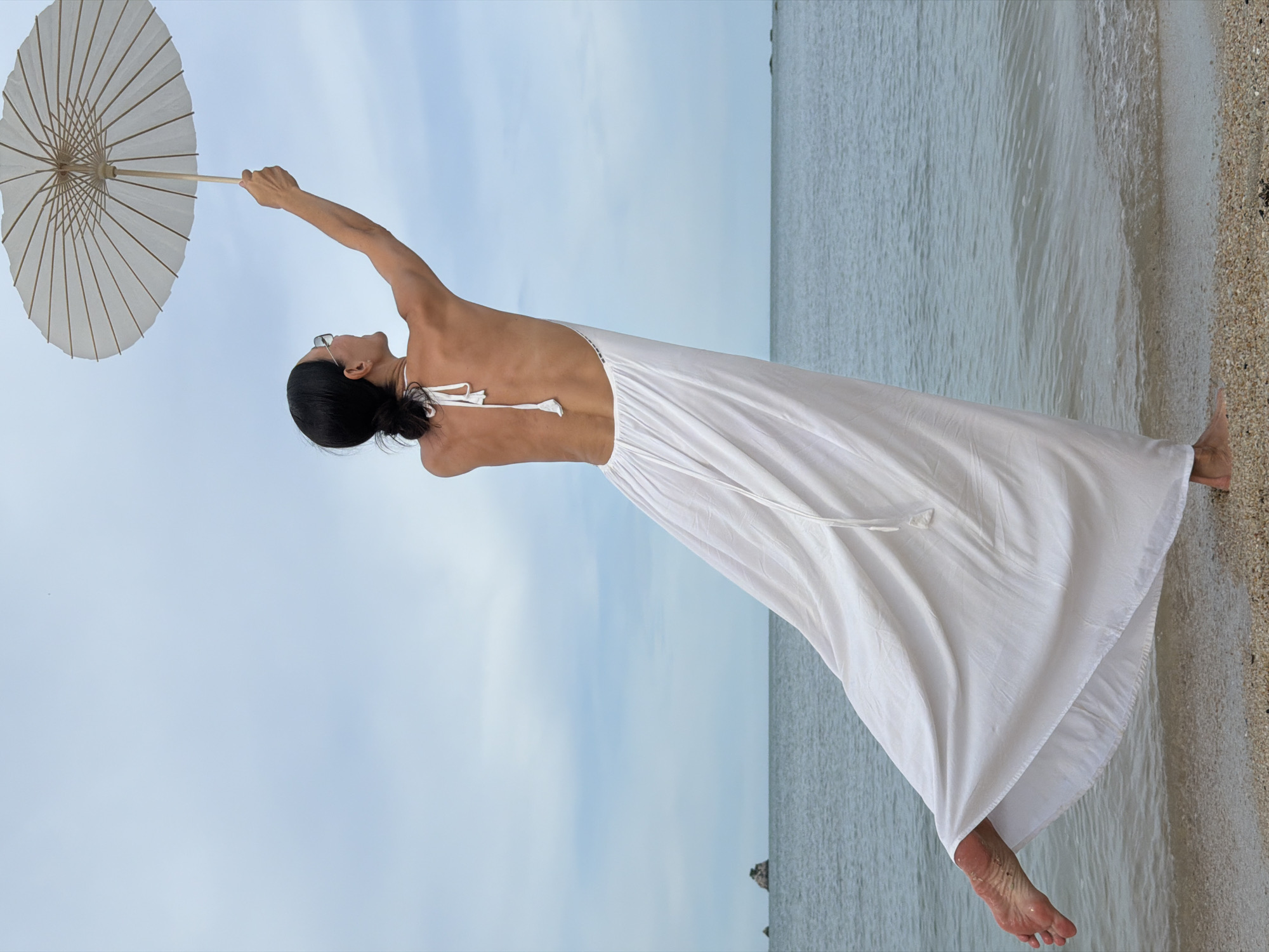 A woman stands on a sandy beach, her body angled away from the camera, and her left arm is raised, holding a white parasol against a cloudy sky. She wears a flowing white dress with a low back and thin straps. Her feet are bare, with the left foot lifted and visible beneath the hem of her dress. The ocean is to her right, with small waves lapping at the shore. In the distance, a small rocky island is visible. The scene is set in Baan Plai Laem, Thailand.