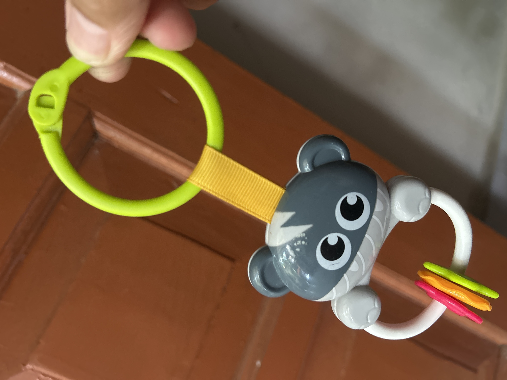 The image shows a close-up of a baby toy hanging against a wooden door. The toy features a grey and white cartoon animal face with large, circular eyes. Below the animal face is a white ring with three colorful, stick-like rattles (pink, orange, and green) attached. A yellow ribbon connects the animal toy to a bright green plastic ring, which is being held by a person's fingers. The background is a dark, reddish-brown wooden surface, likely a door, with some visible grain and texture. The lighting suggests an indoor setting. This image was captured in Playen, Indonesia.