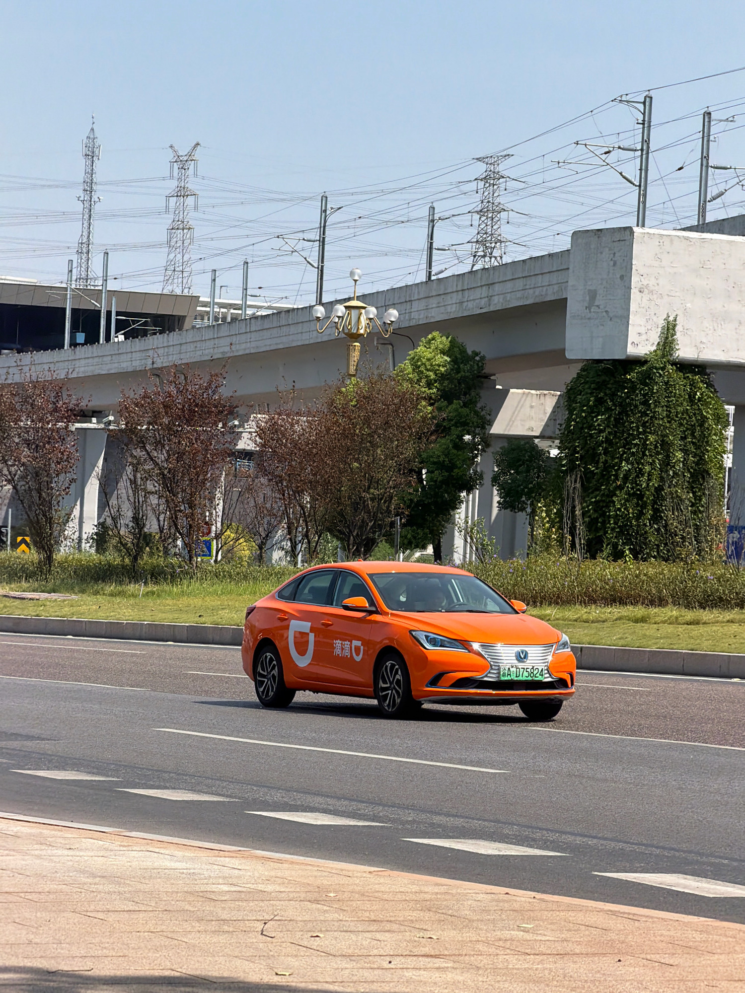 An orange Didi Chuxing ride-hailing vehicle is captured in motion on an asphalt road. The car displays the Didi logo on its side and has a license plate reading "A D75824". In the background, elevated railway tracks are visible, supported by concrete structures. Tall, industrial power line towers and wires crisscross the sky. The environment includes green grass and trees, with some deciduous trees showing brown foliage. The scene appears to be set in a modern urban area, potentially in Jiangjin, China, given the Didi vehicle and the infrastructure. The sky is clear and blue, indicating a sunny day.