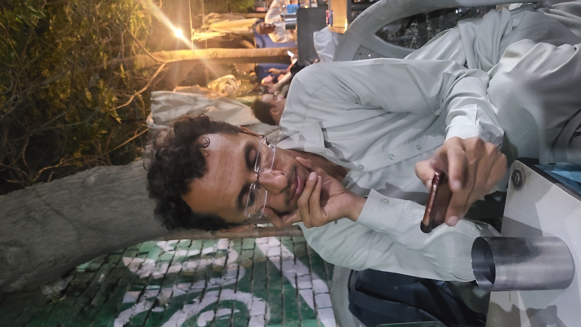 This image captures a nighttime scene in what appears to be a casual outdoor or semi-outdoor setting. The primary subject is a man with dark, curly hair and clear-framed glasses, wearing a light-colored (likely white or light grey) button-up shirt, possibly part of a shalwar kameez. He is lying on his side, resting his head on his left hand, and looking at a brown smartphone held in his right hand. His expression appears calm and focused.
Beside him, on a surface, is a metallic cylindrical object, which could be a cup or container. The ground features a distinctive green and white geometric pattern. In the background, other people are visible in various states of relaxation; one person is seen lying down behind the main subject, and others are further back, some seated.
The scene is illuminated by artificial lights, which create a bright glow and lens flares, confirming it is nighttime. Sparse vegetation or trees are discernible in the dark background. The overall atmosphere suggests a relaxed gathering or waiting area. No visible text is present. The attire and general environment are consistent with a location such as Quetta City Tehsil, Pakistan.