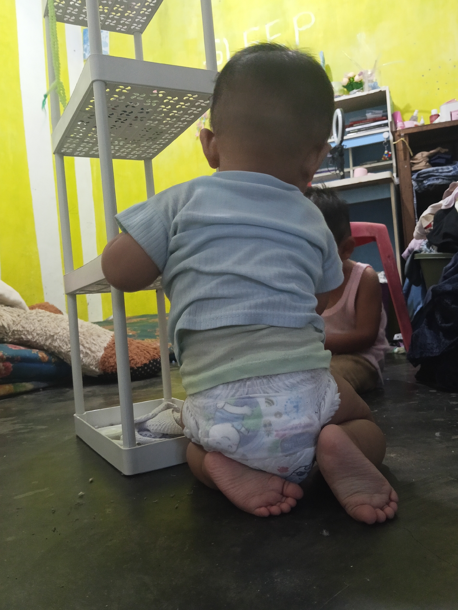 A rear-view, medium shot captures a toddler wearing a light blue t-shirt and a diaper, kneeling on a dark, dusty floor in what appears to be a cluttered room in Lestari Indah, Indonesia. The toddler's head is tilted down, obscuring their face. To the left, a white, tiered plastic shelf unit with a floral-patterned design is positioned. Further back, to the right, another child is partially visible, wearing a pink tank top and light brown pants, seated and facing away from the camera. Behind the seated child are shelves filled with various items, including books, containers, and clothing, suggesting a living space. The wall behind the toddler is painted a bright yellow with faint white lettering that appears to read "DEEP". Pillows and blankets are scattered on the floor to the left of the toddler. The overall environment appears to be an indoor domestic setting, likely a child's play area or bedroom, characterized by a mix of organized storage and scattered personal belongings.
