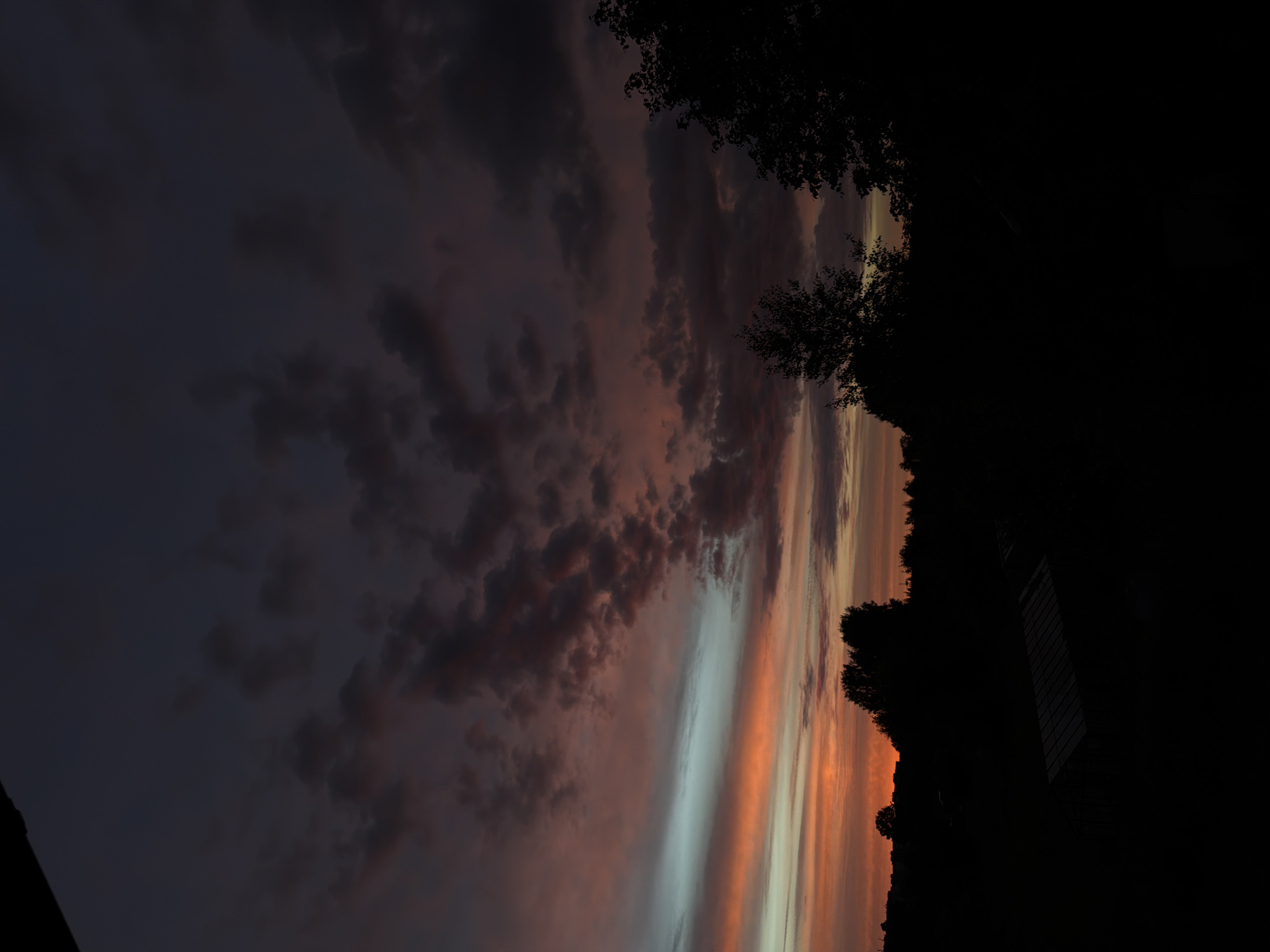 This is a vertical, low-angle, dimly lit photograph of a sunset over an outdoor, rural area. The sky dominates the upper portion of the frame, displaying hues of deep purple, dark gray, and bands of vibrant orange and light blue from the setting sun. Scattered clouds are visible throughout the sky, with some appearing reddish-brown and others dark gray. The lower third of the image consists of a silhouetted landscape. In the foreground, a dark, rectangular structure, possibly a greenhouse or shed with a corrugated roof, is visible. Behind it, the silhouettes of various trees and foliage stretch across the frame, with a more dense grouping of trees on the right side. The background shows a faint outline of distant buildings or structures, suggesting a settlement or village on the horizon. The overall impression is of twilight in a countryside setting.