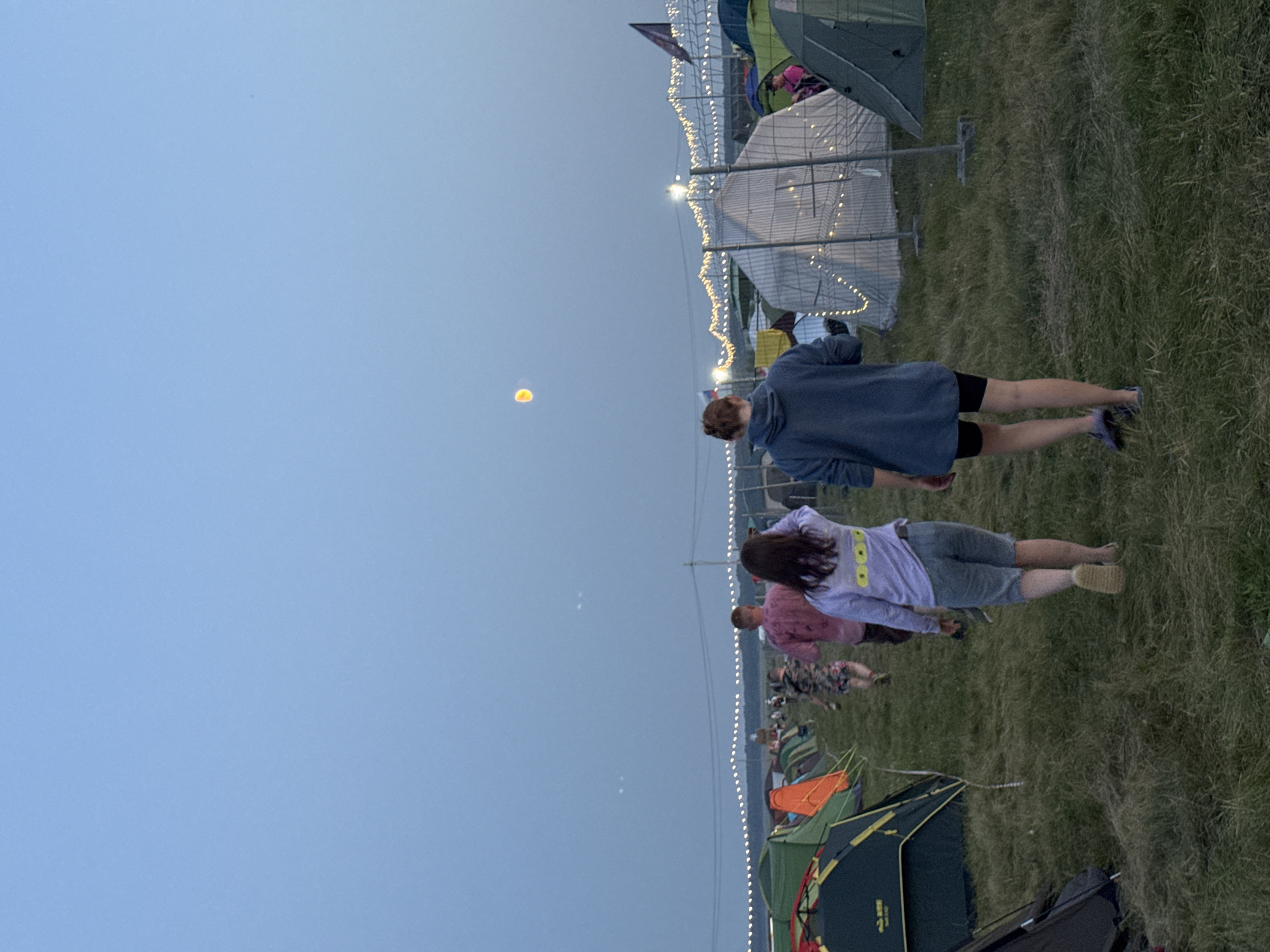 A low-angle, full-shot photograph taken from behind depicts a nighttime festival campsite. Two individuals, one wearing a purple hoodie and shorts and the other a blue hoodie and black shorts, walk away from the camera towards a field filled with tents. Their backs are to the viewer. In the background, additional tents, string lights, and distant figures are visible. The sky is a pale blue with a large, yellow circular object, possibly the moon, and two smaller white orbs. The ground is covered in grass and dry vegetation.