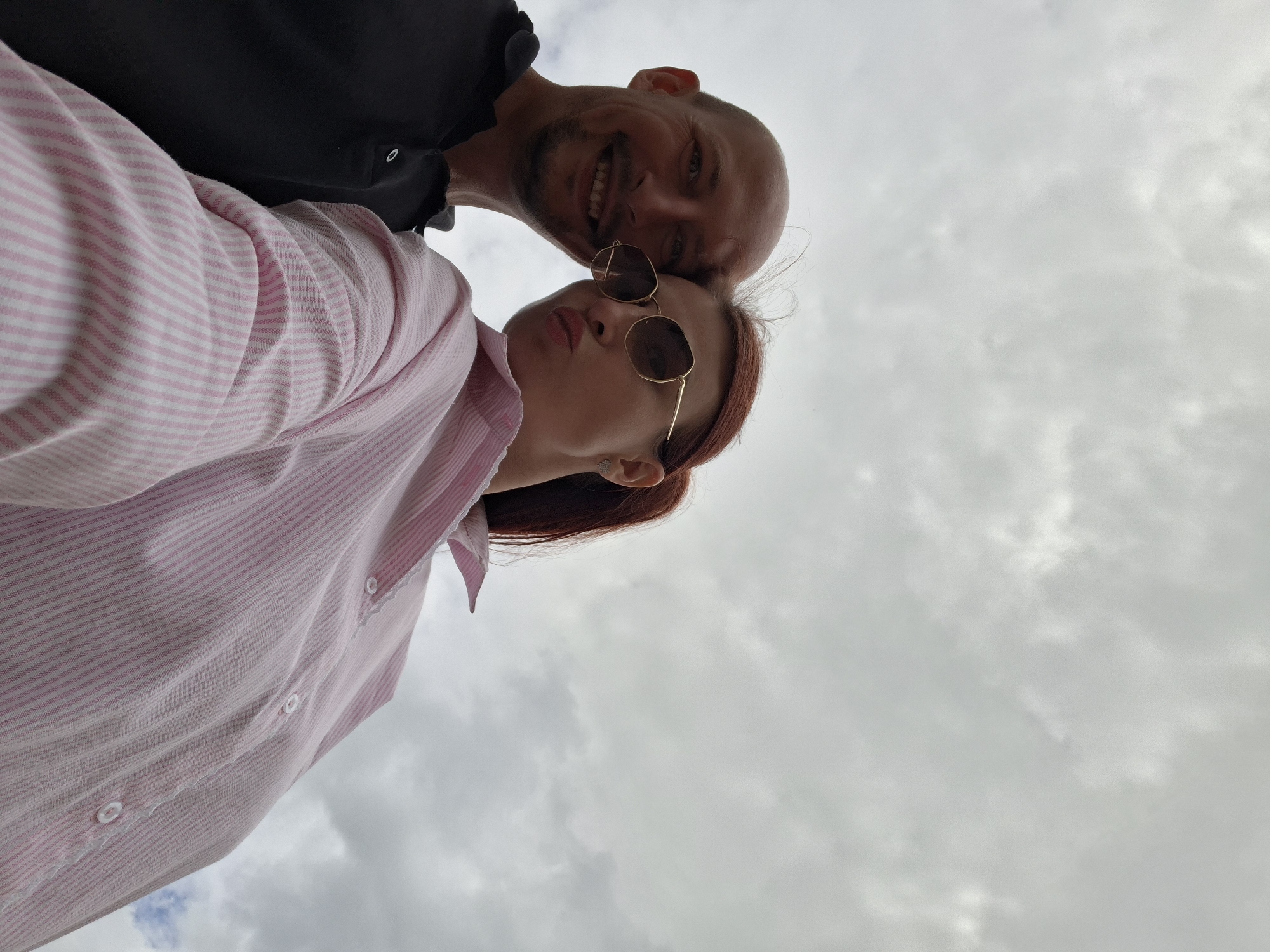 A low-angle, wide shot shows a bald, fair-skinned man with a slight smile and a fair-skinned woman with red hair pulled back into a ponytail, wearing sunglasses and puckering her lips. The man is wearing a black polo shirt, and the woman is wearing a pink and white striped button-down shirt. They are positioned closely together, with the woman's head tilted forward and resting against the man's. The background is a cloudy, grey sky. This image was captured in Myadzyel, Belarus.