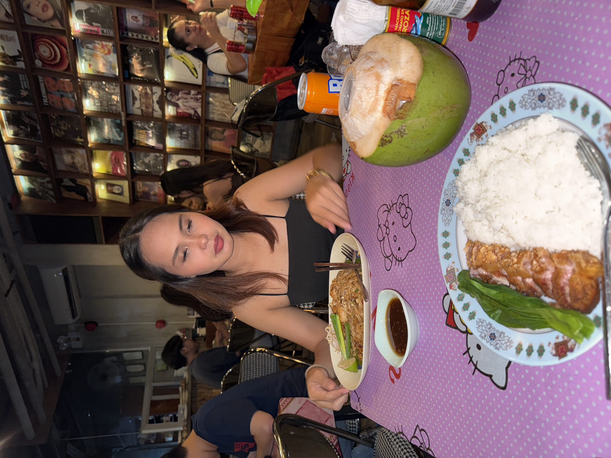 In a restaurant in Davao City, Philippines, a young woman with long, dark hair sits at a table adorned with a pink Hello Kitty-themed tablecloth. She is wearing a black spaghetti strap top and is looking down at a plate of Pad Thai noodles, a lime wedge, and bean sprouts. A small bowl of dipping sauce is placed in front of the plate. To her right, a whole young coconut with its top partially removed rests on the table, along with a can of soda and a bottle of sauce. In the background, a wall is covered with what appear to be framed records or album covers. Other patrons and restaurant staff are visible in the background.