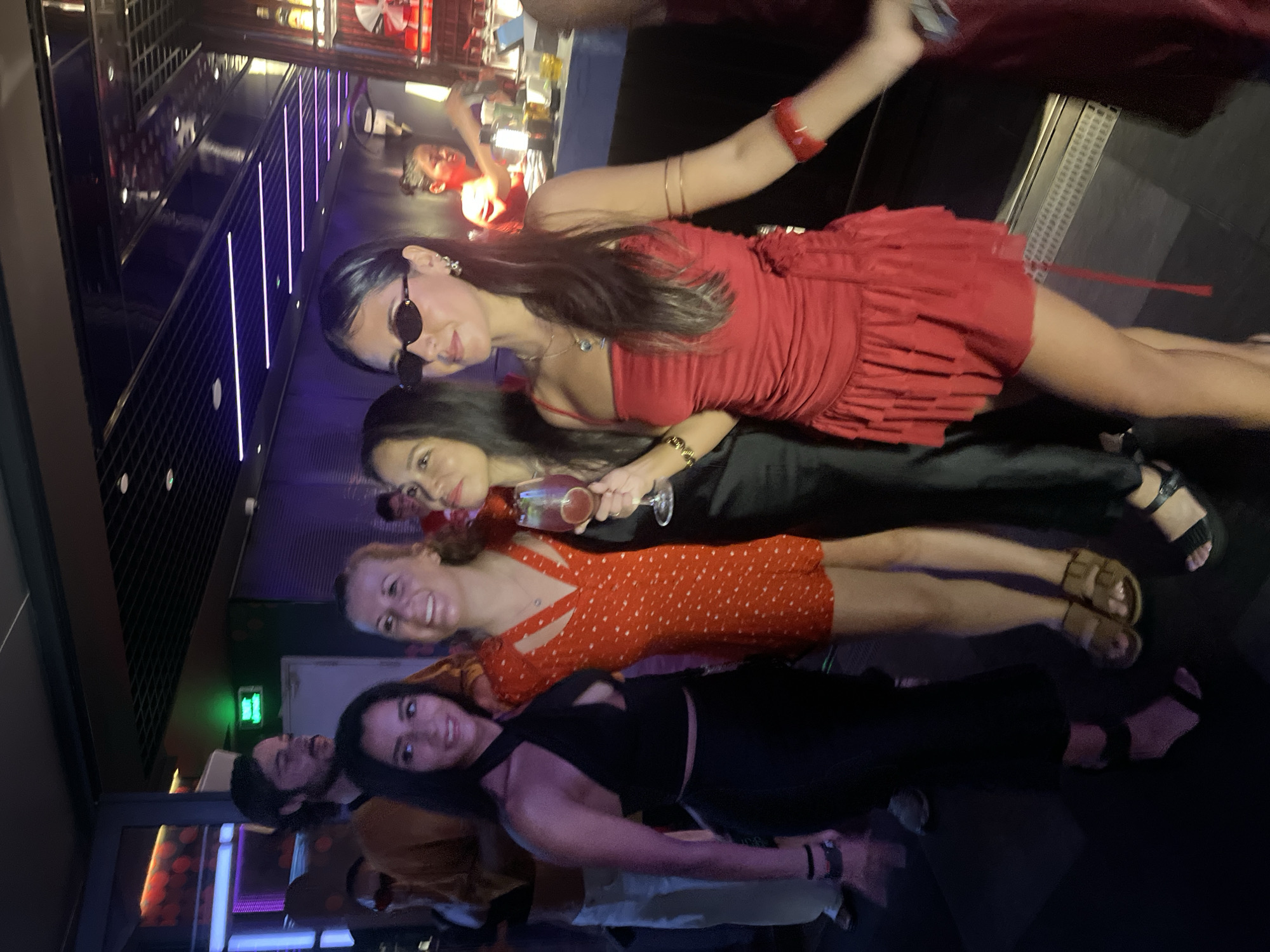 In a dimly lit nightclub, four women and a man are socializing. The women, dressed in festive attire, are standing close together, appearing to be engaged in conversation. The woman on the far left wears a black halter top and skirt, while the woman next to her is in a red and white polka dot dress. The third woman is in a black dress, holding a glass of pink liquid, and the woman on the right is in a red ruffled dress, wearing sunglasses. The man, with dark hair and a beard, stands behind them, partially obscured. The nightclub environment is characterized by purple and pink lighting, a bar in the background, and a dark floor. The scene suggests a lively social gathering in Đà Nẵng, Vietnam, likely at a bar or club.