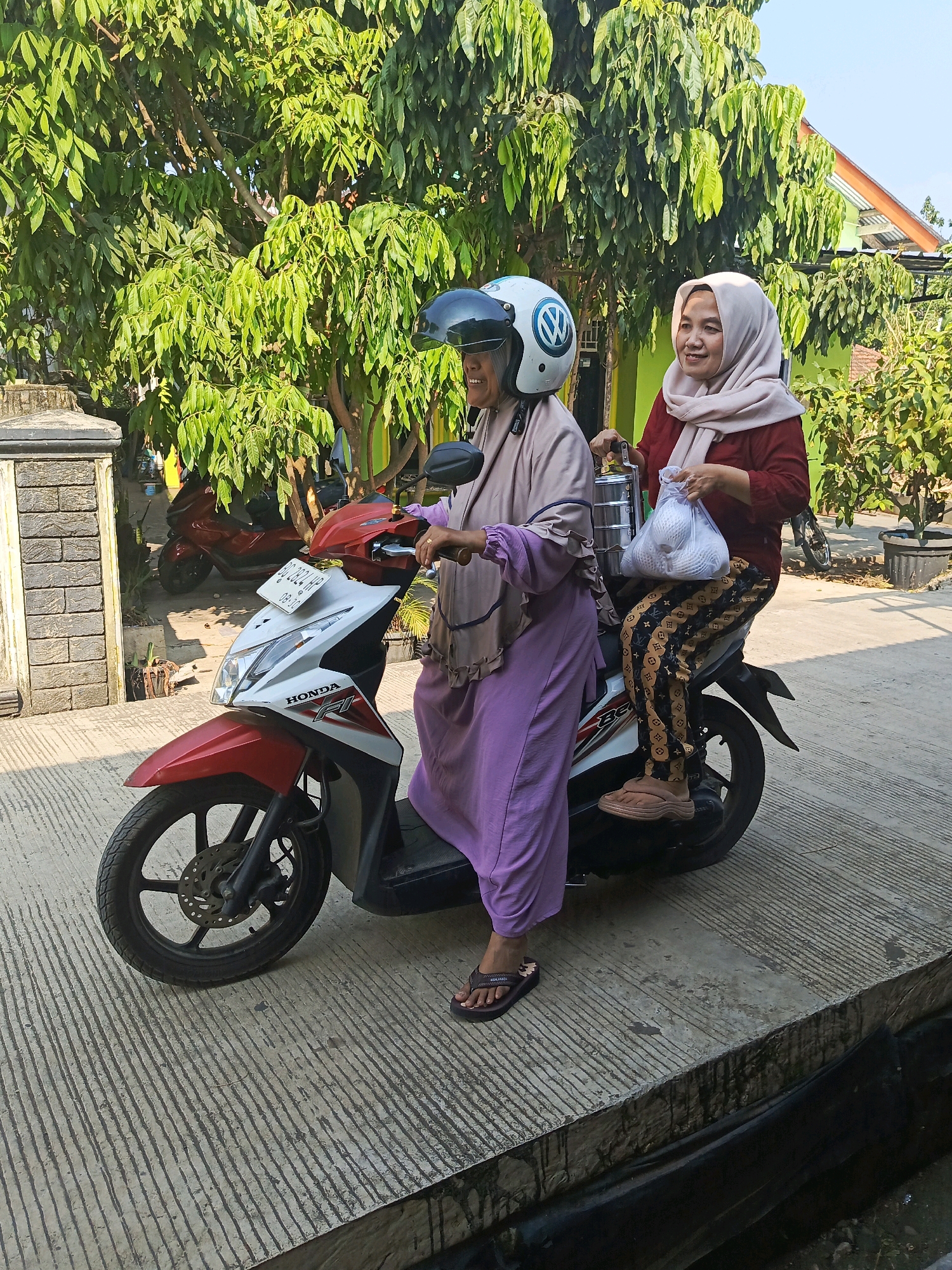 Two women are seated on a red and white Honda scooter. The woman in front is wearing a light purple abaya and a white helmet with a blue Volkswagen logo. She is holding the scooter's handlebars. The woman behind her is wearing a maroon long-sleeved shirt, a light pink hijab, and patterned pants. She is holding a silver tiered food carrier in her left hand and a white plastic bag filled with items in her right hand. Both women appear to be smiling. They are on a paved surface with diagonal lines, possibly a street or driveway, with a concrete curb in the foreground. Behind them, there are trees with lush green leaves and a glimpse of a building with a green wall and an orange-tiled roof. A red scooter is parked in the background. The image is taken in natural daylight, suggesting it is outdoors. The scene appears to be located in Lubuklinggau, Indonesia, given the context provided.