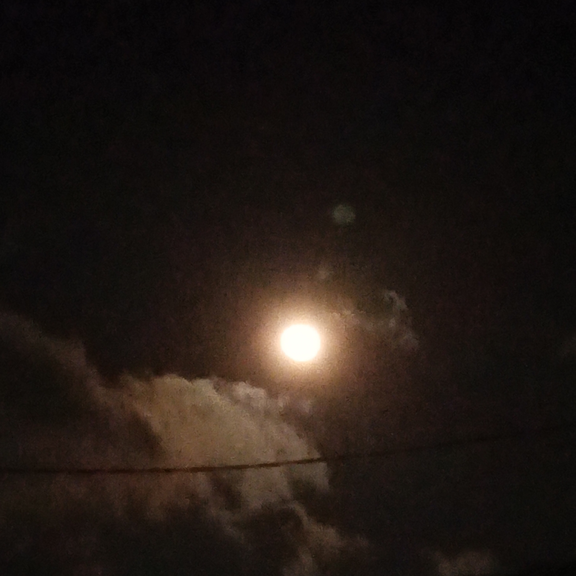 A full moon glows brightly in a dark, cloudy sky, its light illuminating a band of white clouds stretching across the frame. A thin, dark horizontal line, possibly a wire, bisects the image. The overall atmosphere is dimly lit and somewhat obscured by the night and clouds, suggesting a nighttime scene in a location like Cigadung, Indonesia.