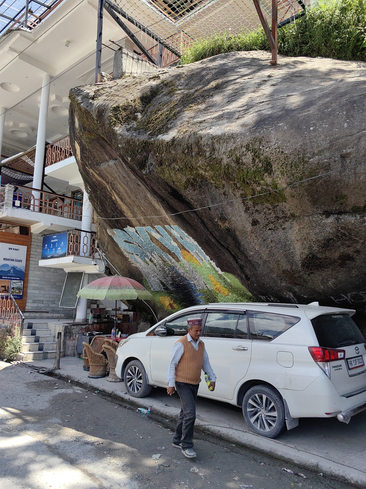 The image depicts a vibrant street scene in Vashisht, India, characterized by a massive, moss-covered rock formation dominating the right side of the frame. A colorful mural, featuring abstract or landscape elements in blue, green, and yellow, is painted on the lower section of the rock. Above the rock, a green safety net is visible, supported by a metal frame.
To the left of the rock stands a multi-story white building, identified by signs as "SKYH! MOUNTAIN ESCAPE" and "BOUTIQUE HAP & ROOFTOP RESORT," indicating a hotel or guesthouse. The building features balconies with brown railings and a translucent roof structure at its apex. Stone stairs lead up to an entrance on the ground level.
Beneath the rock's overhang, a small vendor setup is present, marked by a green and pink striped umbrella. Several light brown wicker chairs and a partial table are visible, alongside various bottles and two dark chalkboards, suggesting a food or beverage stall.
A white Toyota Innova Crysta Multi-Utility Vehicle (MUV) is parked parallel to the curb, facing left. Its rear license plate reads "HR 13F 0029," and a "SATYAM TOURS" sticker is affixed to the rear window.
A single man is walking in the foreground, from right to left, passing the parked car. He wears a maroon and white patterned cap, a light blue collared shirt, a brown knitted sleeveless sweater, dark trousers, and sneakers. He carries a green bottle in his left hand.
The paved road is uneven and shows signs of wear, with a raised curb separating it from a narrow, debris-strewn edge where a small drainage channel is visible. Shadows from overhead structures or foliage are cast across the ground, indicating daylight conditions.