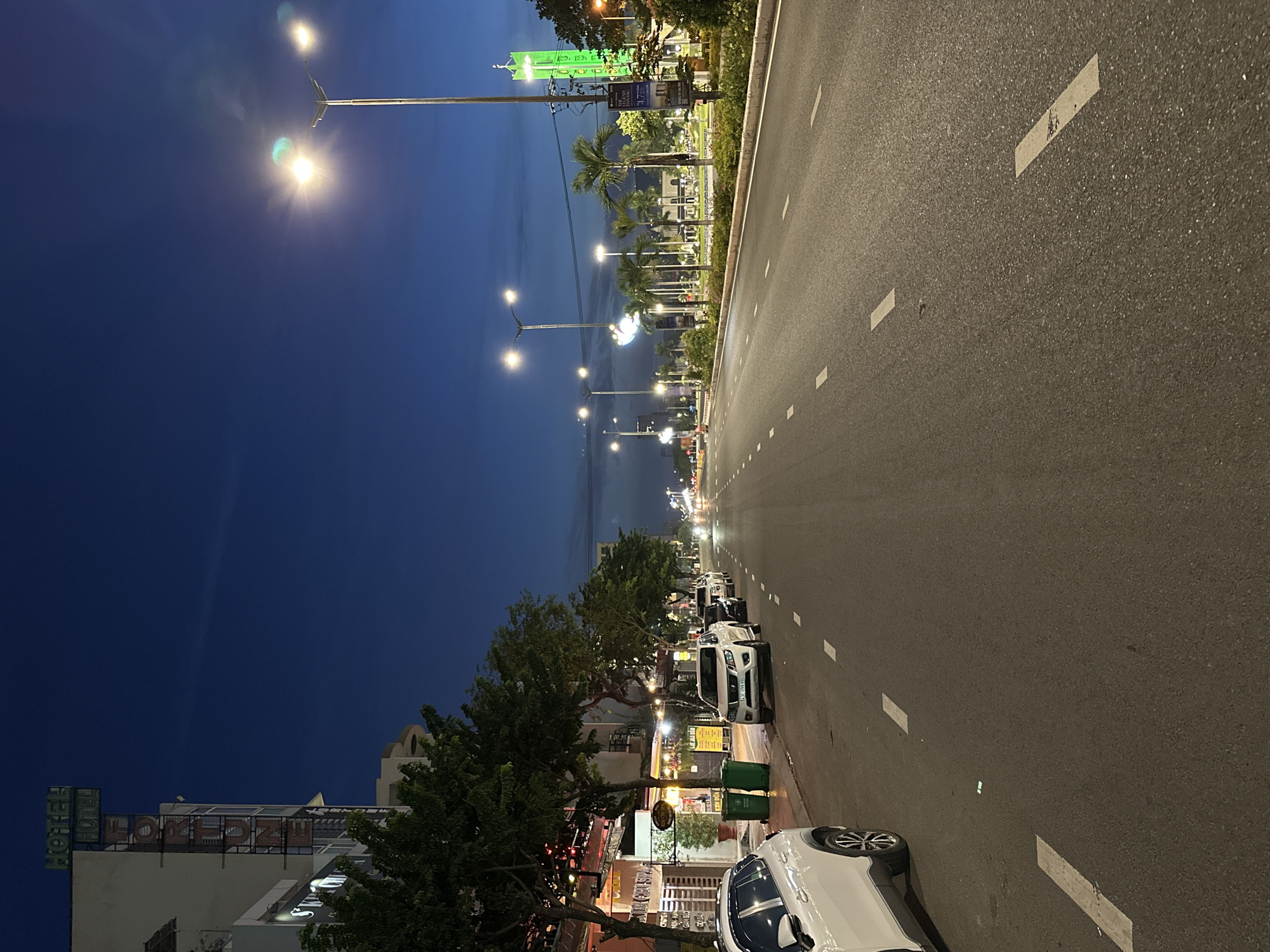 This is a night-time, eye-level, medium shot of a wide, paved road in Da Nang, Vietnam. The road is illuminated by streetlights, and several cars are parked along the right side. The left side of the road features a sidewalk with trees and buildings, including a building with a sign that reads "HOTEL FORTUNE." The sky is a deep blue, with some faint clouds visible. The overall scene is peaceful and quiet, suggesting a late hour.