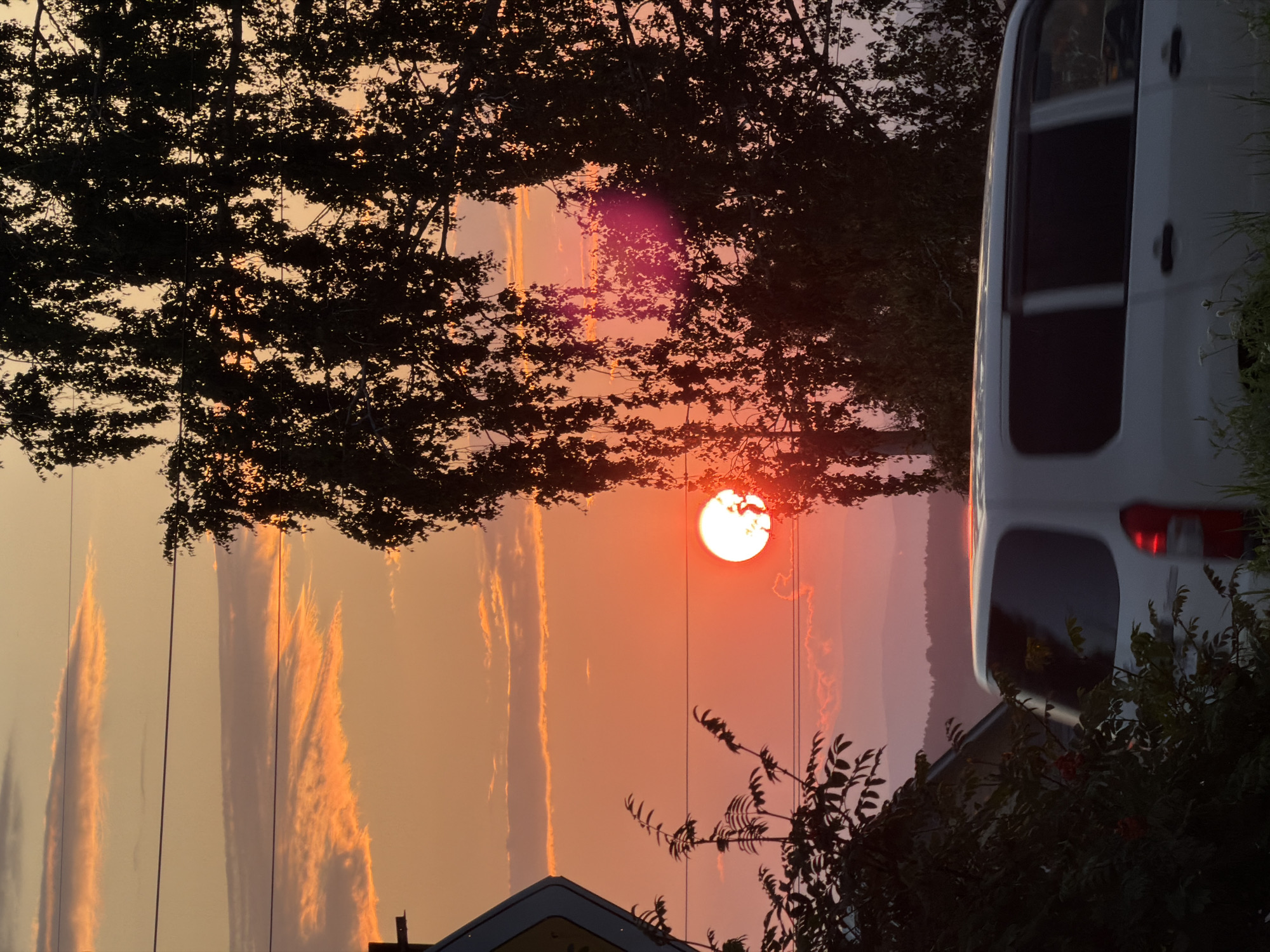 The image captures a sunset scene, with the sun prominently displayed as a bright, glowing orb just above the horizon. The sky is a gradient of warm colors, transitioning from orange and yellow near the sun to a softer pink and purple further up. Wispy clouds are scattered across the sky, catching the sunset light. In the foreground, a silhouette of a large tree with dense foliage partially obscures the view of the sky. To the left, a portion of a road sign and some greenery are visible. A white van is seen driving in the lower right portion of the image, its rear taillight emitting a red glow, suggesting motion blur. The background reveals a hazy, layered landscape of distant hills or mountains, fading into the twilight. The overall impression is one of a serene, natural landscape bathed in the warm light of dusk, with a touch of human presence introduced by the road sign and the moving vehicle.