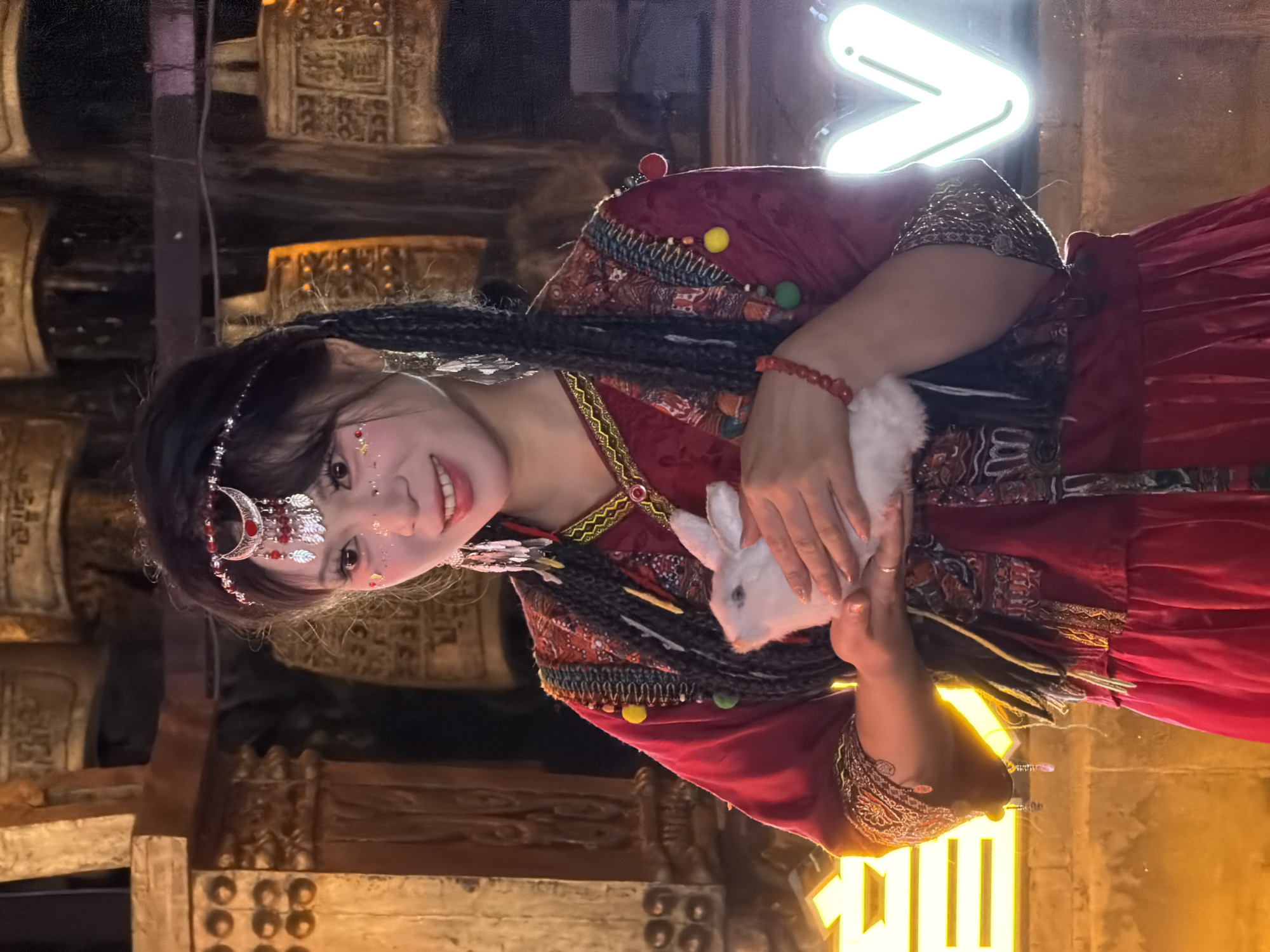 A young woman with dark braided hair and festive makeup, including glitter on her cheeks and eyelids, stands outdoors at night. She is wearing a red traditional Chinese dress adorned with colorful embroidery and beaded trim. In her hands, she holds a small, fluffy white rabbit. The background features a display of large, ornate brass bells and a bright neon "V" sign, suggesting a cultural performance or festival setting in Yuzhong District, China. Yellow neon lights are also visible on the left side of the image.