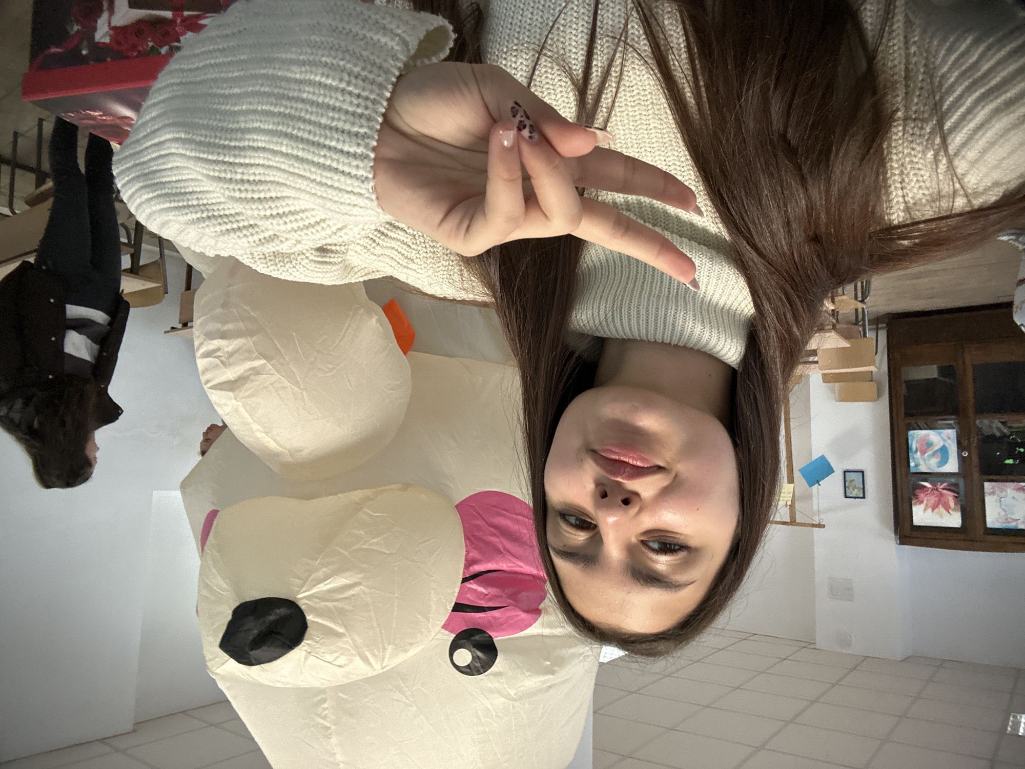 A young woman is posing for a selfie indoors, making a peace sign with her right hand. She is wearing a cream-colored, chunky knit sweater. Long, dark hair falls down her shoulder and side of her face. Next to her and mostly visible behind her is an inflated polar bear decoration with a black nose and a pink tongue. Two other people are partially visible on the left side of the frame. The location appears to be inside a room with white walls and tiled floors. In the background on the right, there is a dark wood cabinet with pictures or paintings inside. Small framed pictures hang on the wall. The location is Panagyurishte, Bulgaria.
