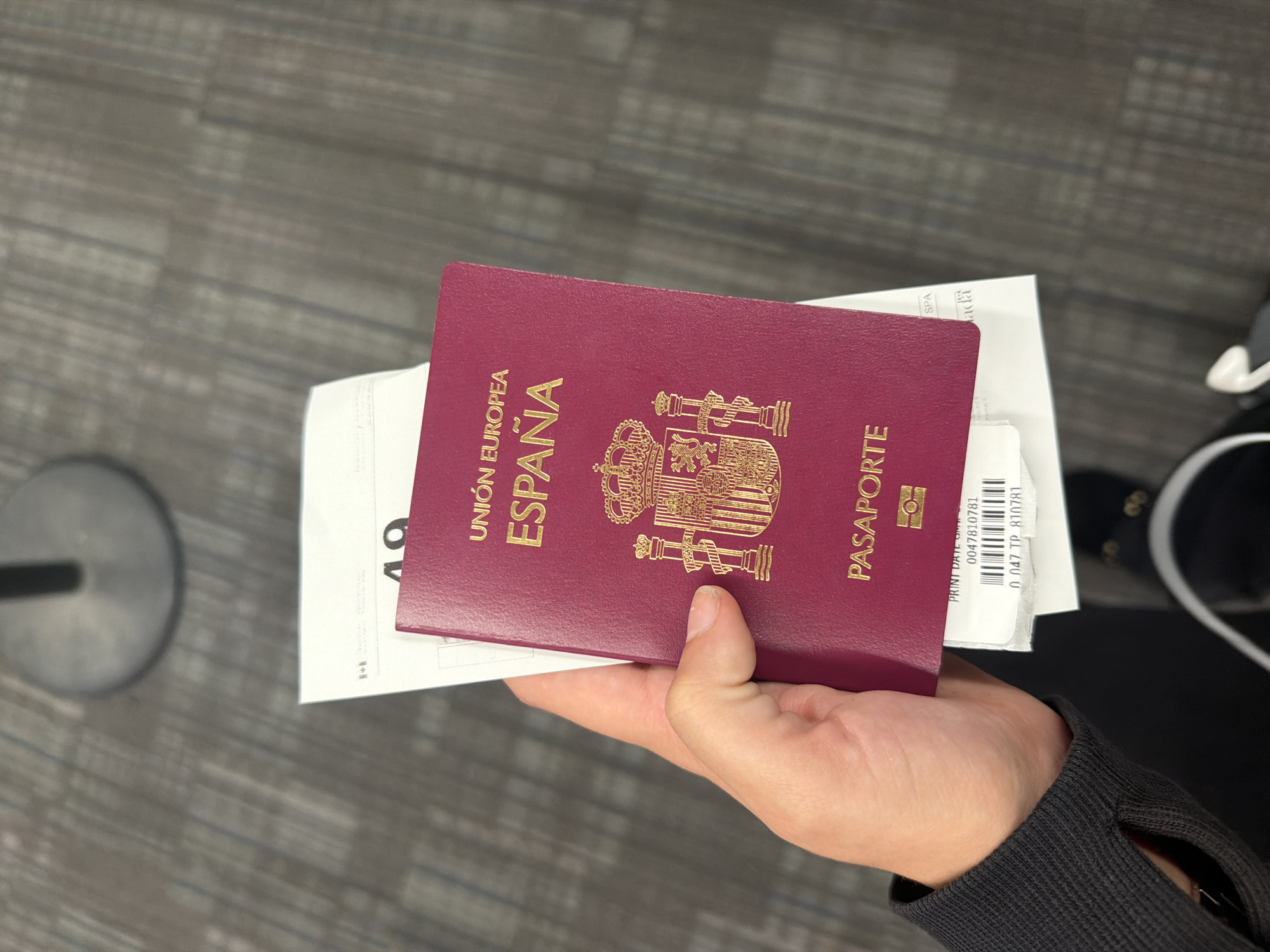 A person's left hand holds a maroon-colored Spanish passport and white paper travel documents in a downward-angled view. The passport prominently displays "UNIÓN EUROPEA," "ESPAÑA," the Spanish coat of arms, and "PASAPORTE" in gold embossing. Partially visible beneath the passport are several white paper documents. One document shows a large black number "49" and a faint Canadian flag symbol with the word "CANADA" partially visible. Another document reveals "PRINT DATE" followed by "0047810781," a barcode, and "0 047 TP 810781." The letters "SPA" are also discernible on one of the documents. The hand is covered by a black, ribbed, long-sleeved garment. The background consists of a textured, gray-patterned floor, characteristic of an airport terminal. A circular, metallic stanchion base is visible out of focus in the upper background. Based on the Canadian flag symbol and "Canada" visible on the travel documents, along with the airport environment, the location is identified as **Toronto Pearson International Airport (YYZ) in Mississauga, Canada**.