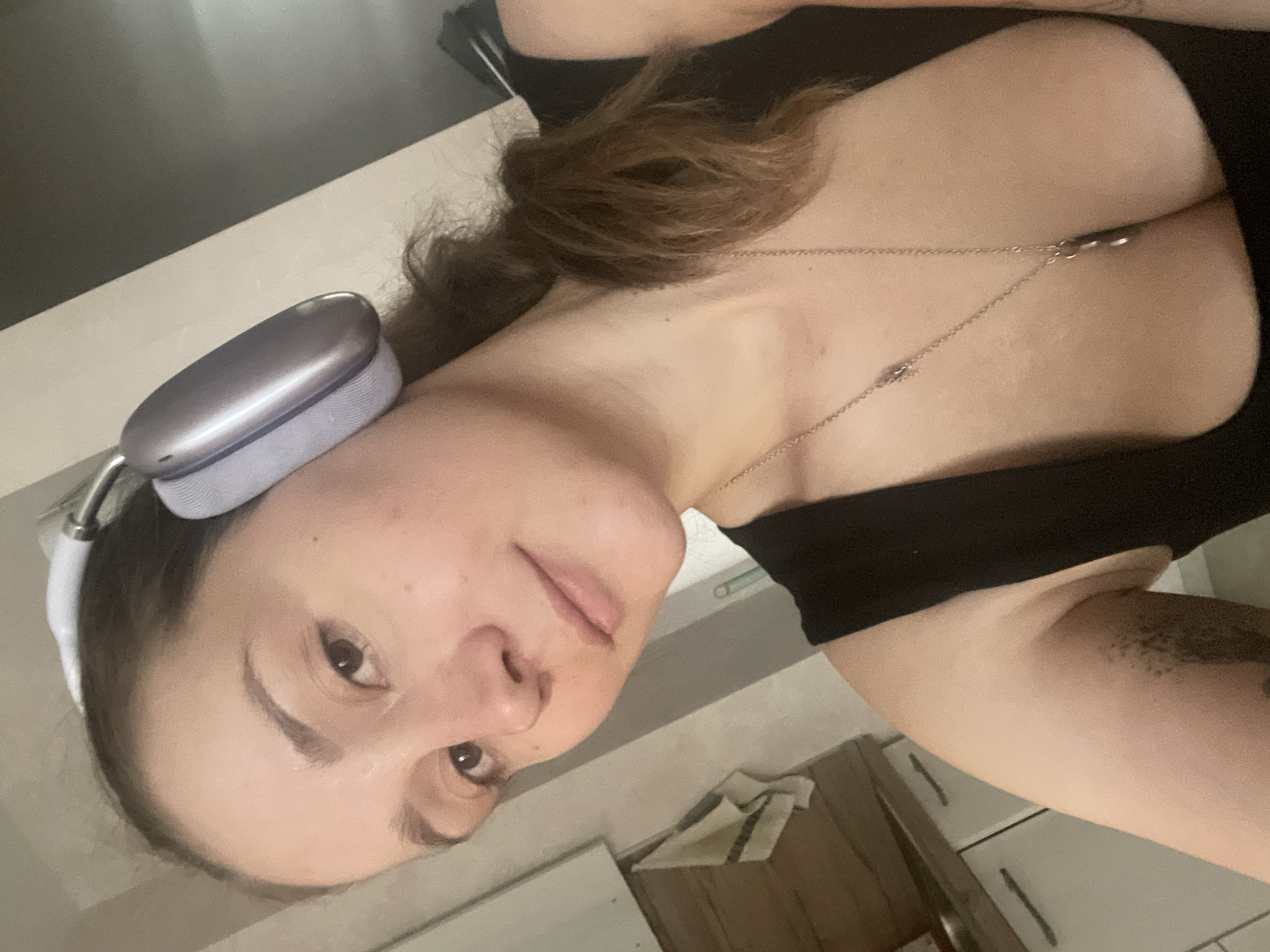 The media file is a selfie taken by a young woman with brown hair. She is wearing a black top and a silver necklace. She has headphones on her head. The background appears to be a domestic setting with cabinets and a counter visible. The lighting suggests it is daytime. There is no visible text in the image.