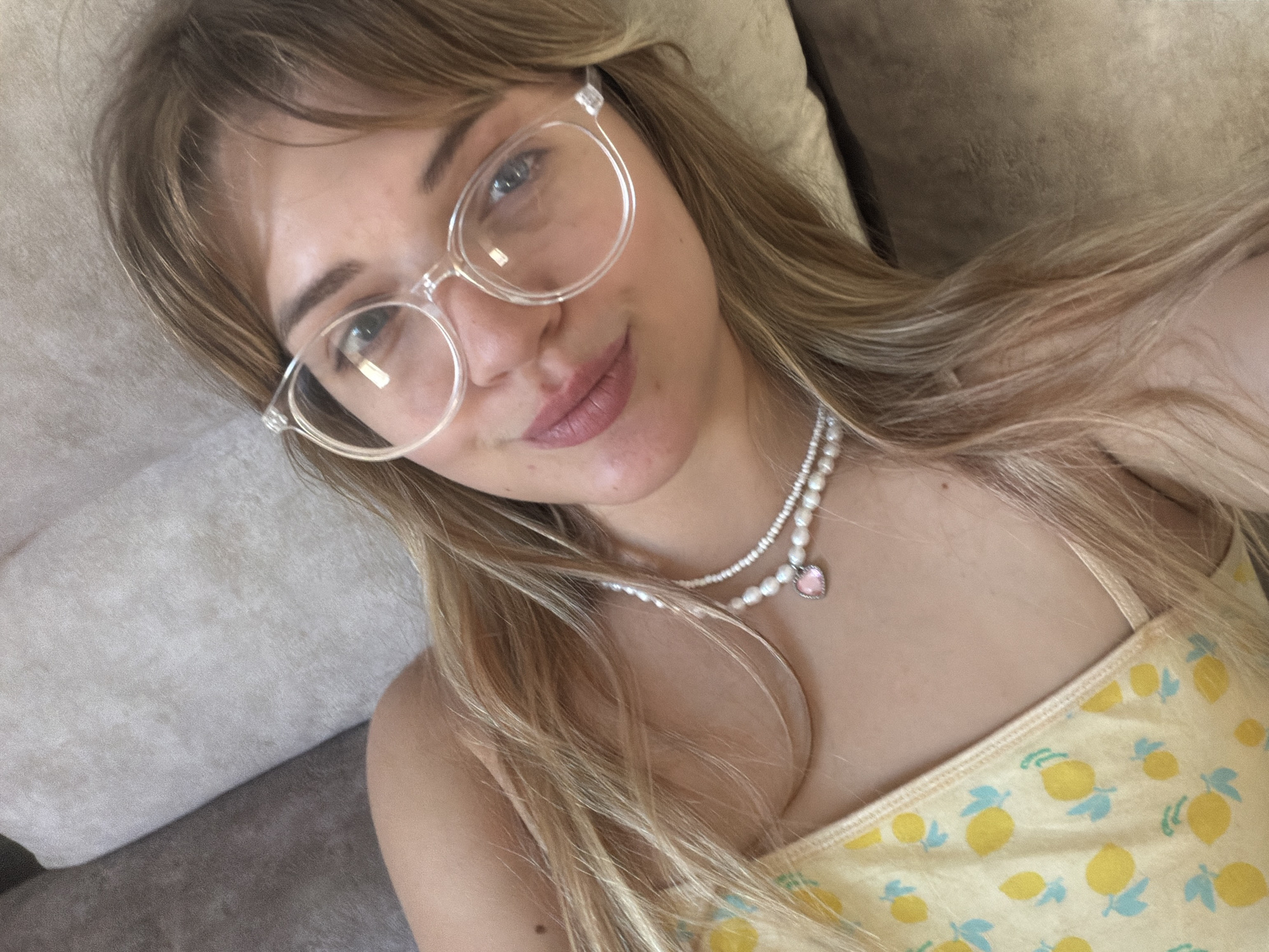 A young woman is lying on a gray upholstered couch or sofa. She is wearing clear-framed eyeglasses, two strands of pearl necklaces (one with a heart pendant), and a yellow top patterned with lemons. She is fair-skinned with blonde hair. The scene appears to be indoors. There is no information to determine if the photo was taken in Большая Шильна, Russia.