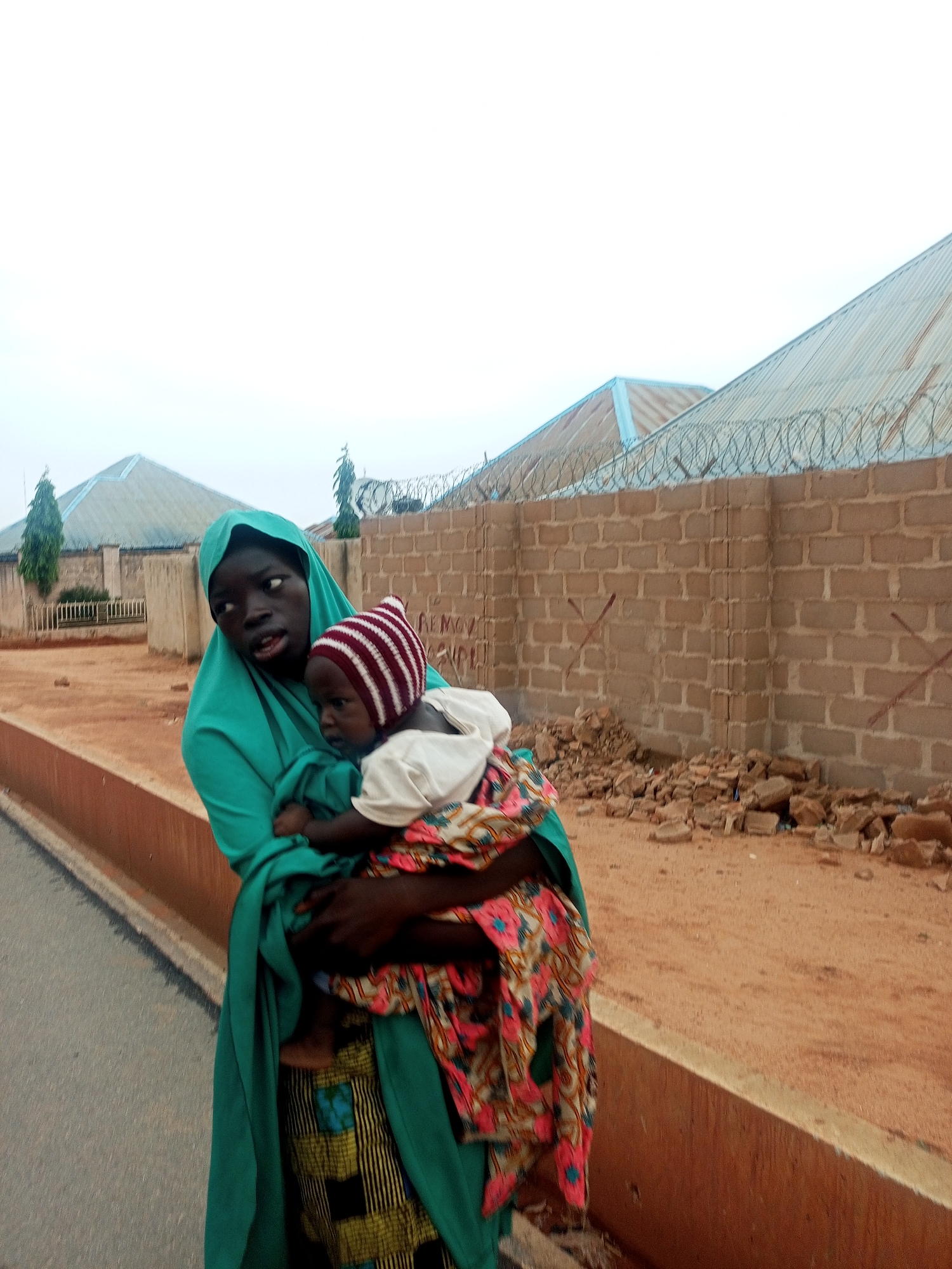 A woman in a green headscarf and patterned skirt holds a baby wrapped in colorful fabric and wearing a striped hat. They stand beside a paved road. Behind them, a brick wall topped with barbed wire sits on a sandy surface littered with rubble. Buildings with corrugated metal roofs are visible beyond the wall. The location is Tempure, Nigeria.