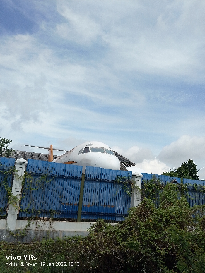 An outdoor, eye-level shot captures the front section of a white airplane, including its cockpit and part of its fuselage, seemingly integrated into a building or structure. The tail fin of the aircraft is also visible behind and above the main body. The airplane appears to be situated behind a tall blue fence made of corrugated metal, which is supported by white stone pillars. The fence itself is partially overgrown with green vines and other vegetation, particularly in the foreground and along its base.
The sky above is partly cloudy with patches of blue, suggesting daytime. No people are visible in the image. The scene suggests an unusual sight, where a large aircraft component is used in an unconventional manner, possibly as part of an attraction or unique dwelling. The overall impression is one of curiosity, presenting a striking contrast between the industrial object and its more natural, overgrown surroundings.
In the bottom left corner of the image, visible text indicates the camera model "vivo Y19s" and a timestamp with names: "Akhtar & Arvan 19 Jan 2025, 10.13". The location of this scene is Sindang, Indonesia.