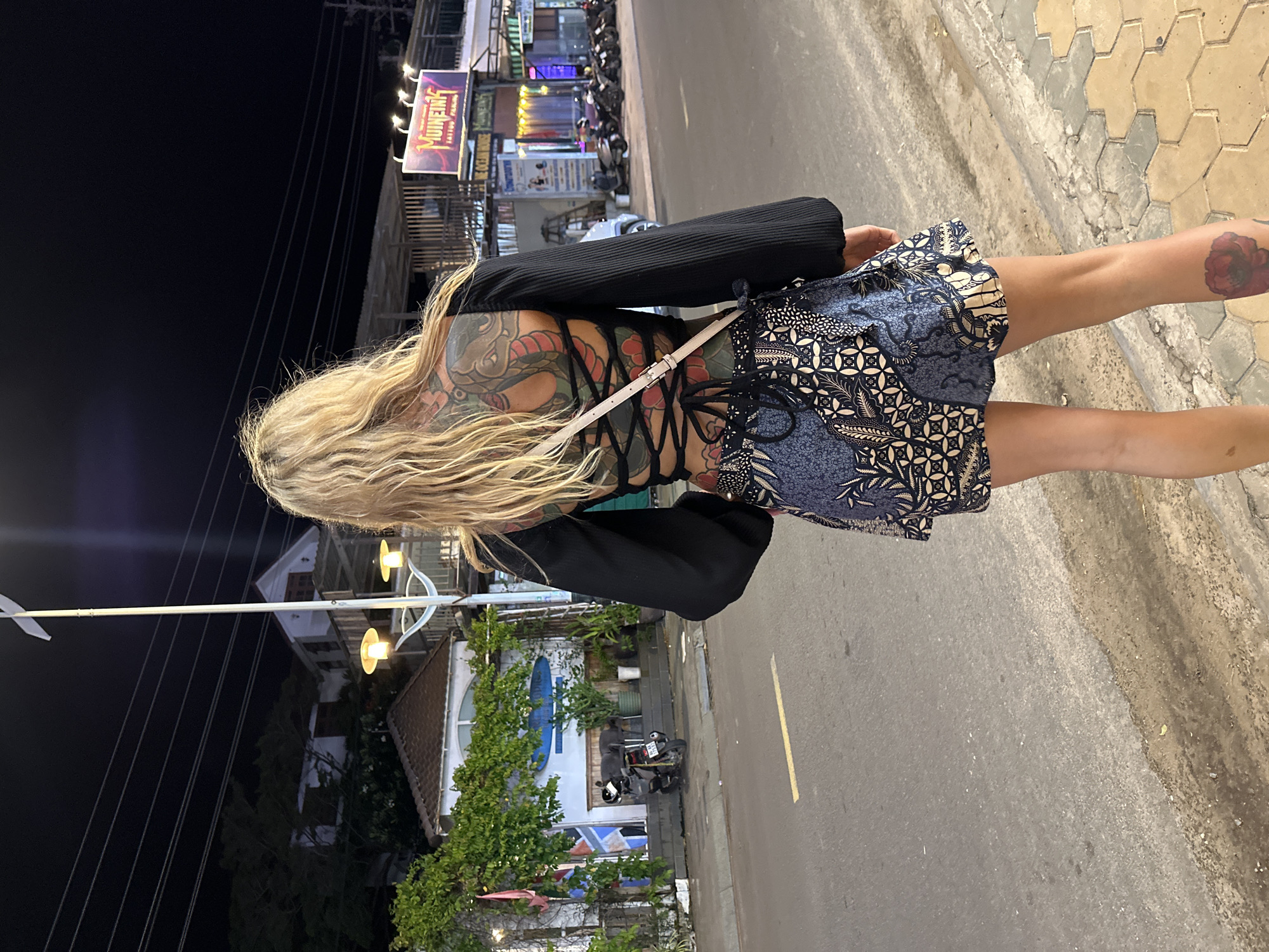 A woman with extensive back and leg tattoos is standing on a sidewalk at night in Phu Thuy Ward, Vietnam. She is wearing a black, open-back, lace-up top and a blue and white patterned skirt. She is facing away from the camera, looking down the street. In the background, there are buildings with neon signs, streetlights, and parked motorcycles. The street is paved, and the sidewalk is made of hexagonal tiles. The scene appears to be a commercial area.
