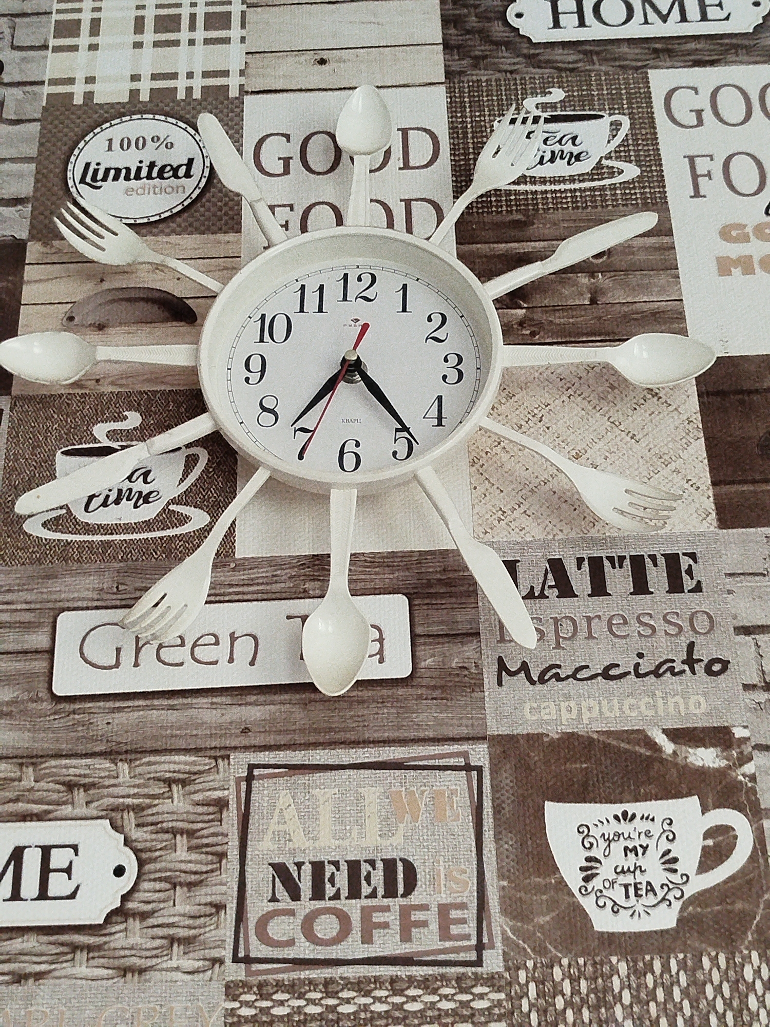 The image shows a close-up of a wall clock with cutlery as hour markers, set against a collage of coffee-themed graphics and textures. The clock face is white with black numbers and hands. Cutlery items such as spoons, forks, and knives are arranged around the clock face to represent the hours. The background features various textures and text, including images of coffee cups, plaid patterns, and words like "HOME," "GOOD FOOD," "TEA TIME," "Green Tea," "LATTE," "Espresso," "Macchiato," and "ALL WE NEED IS COFFEE." A graphic of a teacup with the text "You're my cup of tea" is also visible. The overall impression is of a decorative item for a kitchen or dining area. The image does not contain any information that could indicate its location in Крывасельскі сельскі Савет, Belarus.