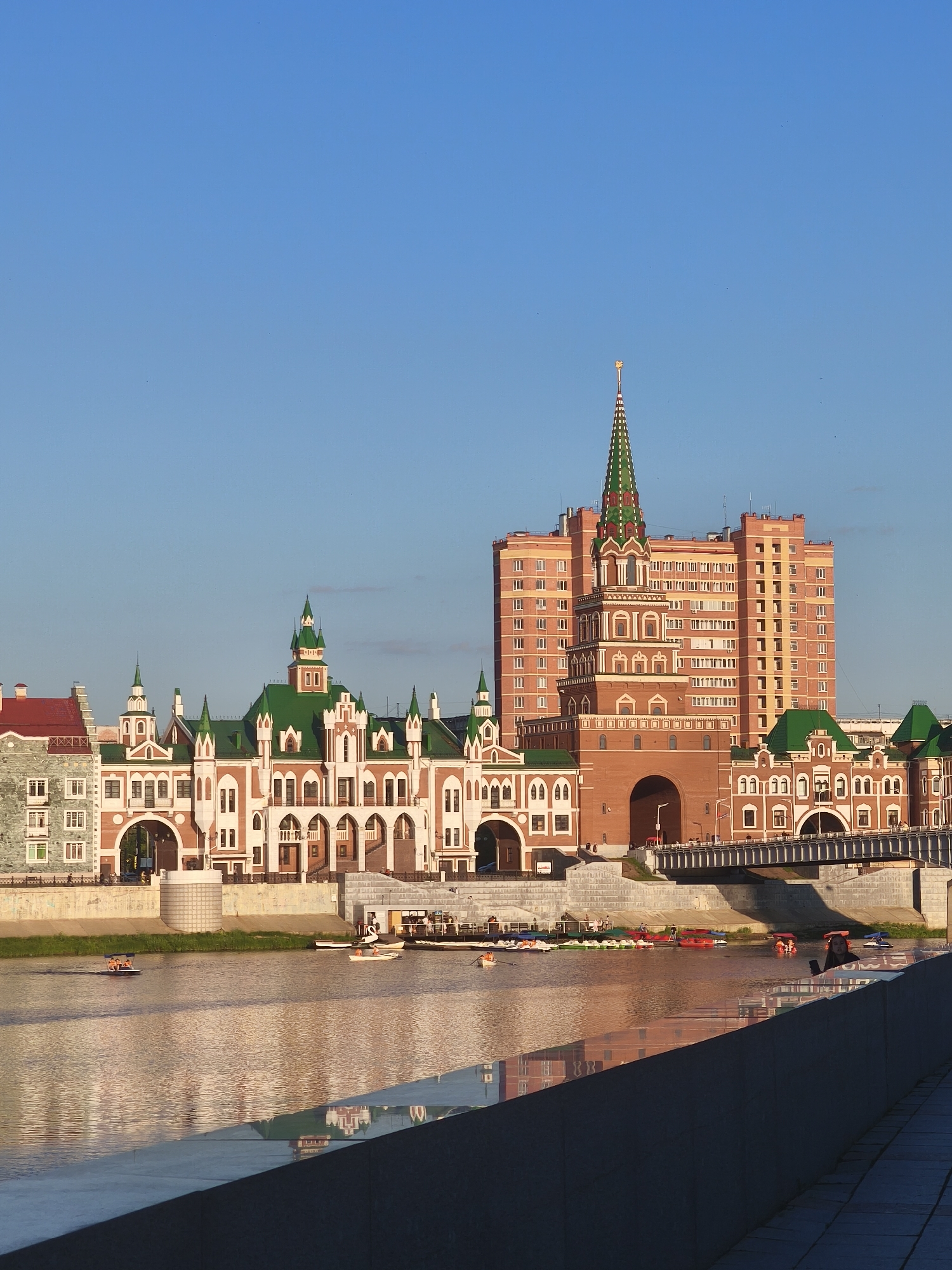 This is an eye-level, full shot photograph of a waterfront scene in Yoshkar-Ola, Russia, featuring the Bruyant Embankment and buildings in a Russian Revival architectural style. The sky is clear and blue, with a few faint birds or insects visible. The sun is shining, casting reflections on the water.
On the left side of the image, a complex of buildings with red-and-white facades and green, pointed roofs dominates the scene. These buildings feature numerous arched windows and doorways, and several small towers. To the right of this complex, a tall, red-brick building with a green, spire-topped tower rises, reminiscent of Kremlin architecture. This building has many windows and appears to be a residential or office building. A bridge spans the river in front of these buildings.
The river in the foreground is calm, with gentle ripples. Several small boats, including pedal boats and what appear to be small motorboats, are scattered across the water. Some people are visible in these boats. On the far right, a portion of a gray, concrete embankment is visible, with a tiled pathway.
The lighting suggests it is late afternoon or early morning, with the sun low in the sky, illuminating the buildings and creating reflections on the water. The overall atmosphere is peaceful and scenic.