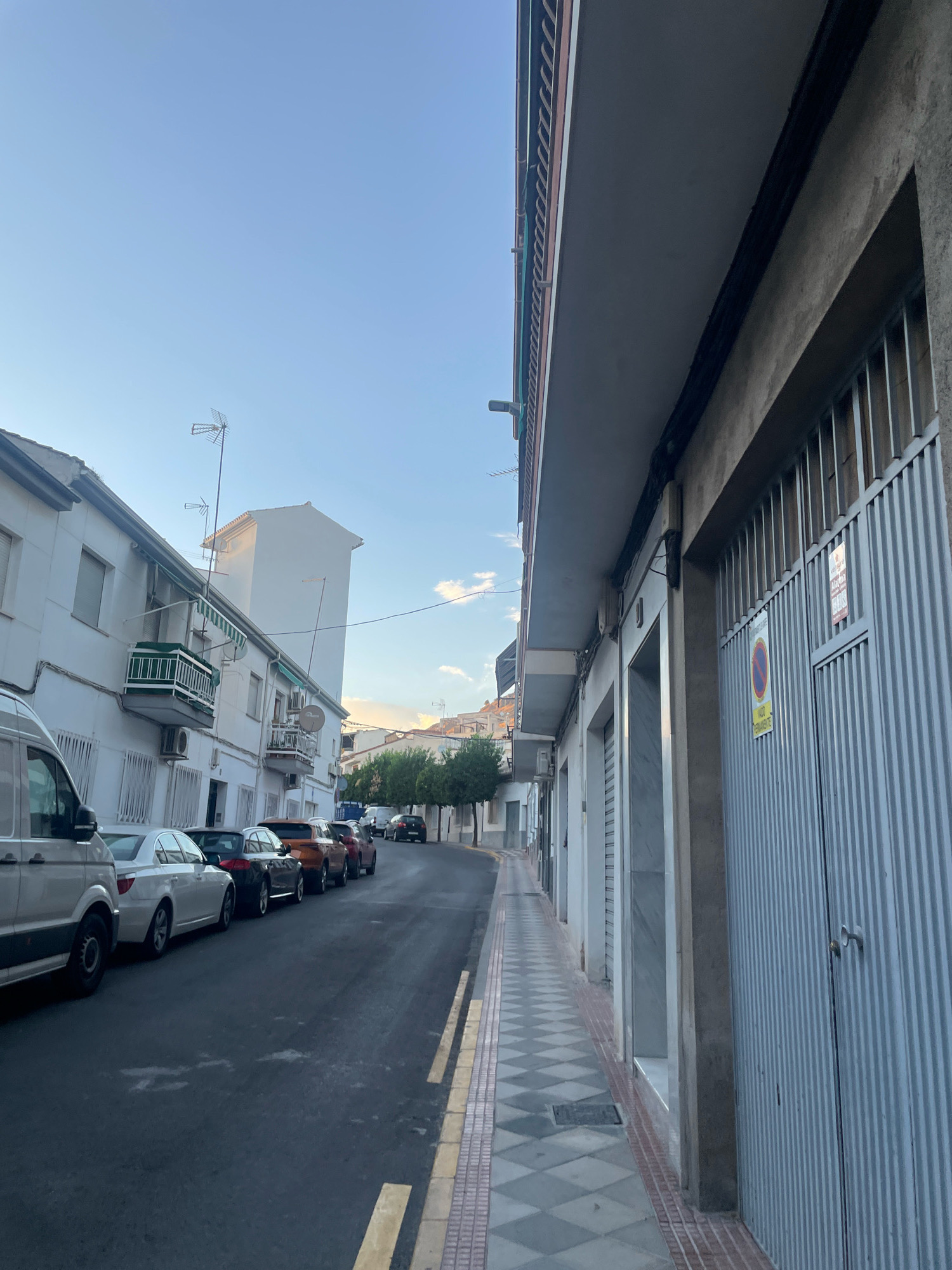 The image depicts a street scene in Atarfe, Spain, likely during the late afternoon or early evening. The perspective is from a low angle, looking down a residential street lined with buildings on both sides.
On the left side of the street, white apartment buildings with balconies and windows are visible. Several cars are parked along the curb, including a white van at the far left, a white sedan, a dark sedan, an orange SUV, and other sedans. A few trees are planted along the sidewalk.
On the right side of the street, a building with a large, corrugated metal gate is prominent. The gate is closed and appears to be a garage or commercial entrance. Two signs are affixed to the gate: one circular with a red border and a diagonal line, indicating a prohibition, and a yellow rectangular sign below it with text. The building also has several closed storefronts with marble-like facades. The sidewalk on this side has a checkered pattern of grey and white tiles, bordered by red bricks.
The sky is a clear blue, with a hint of golden light near the horizon, suggesting either sunrise or sunset. Antennas are visible on top of some of the buildings. The street pavement is dark asphalt. The overall impression is a quiet, ordinary urban street.