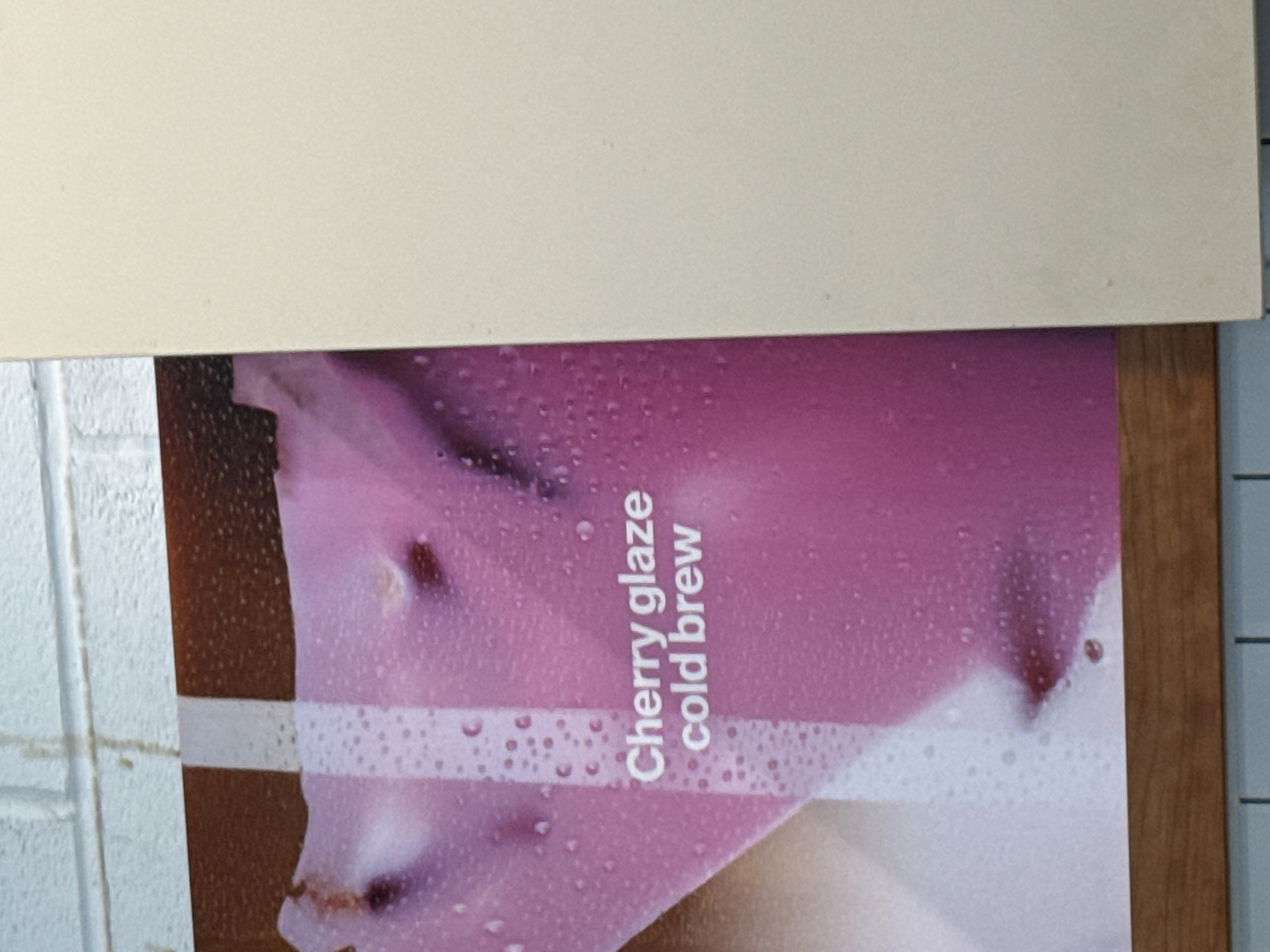 A commercial advertisement poster is affixed to a textured, light-colored wall with visible grout lines. The poster, which depicts a vibrant pink/magenta substance with numerous simulated water droplets and darker reddish-brown inclusions, advertises "Cherry glaze cold brew" in a light-colored font. A horizontal brown wooden strip frames the bottom edge of the poster. A large, plain, light-colored rectangular panel or surface partially obscures the right side of the poster. No individuals are present, and no dynamic actions are occurring. The scene is located in London, United Kingdom.