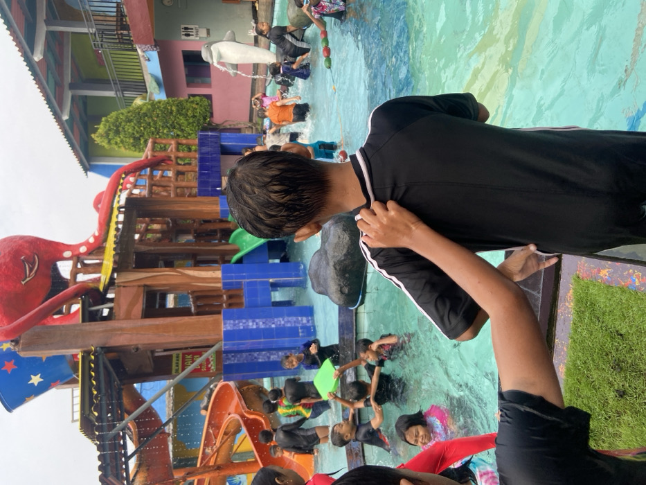 The image shows a waterpark in Kecamatan Tanggulangin, Indonesia. The image was taken at the location (latitude: -7.497, longitude: 112.684). It appears to be a sunny day, and the pool is filled with water. The image captures a boy in the center wearing a black swim suit with white stripes. The boy is being touched on the back by another person, whose arm appears in the image. The boy is looking towards the right side of the image. There are several other children and adults in the waterpark, including a group of children in the water, a group of people standing near the slide, and an adult holding a green rectangular float in the water. The waterpark is decorated with a brightly colored octopus sculpture.