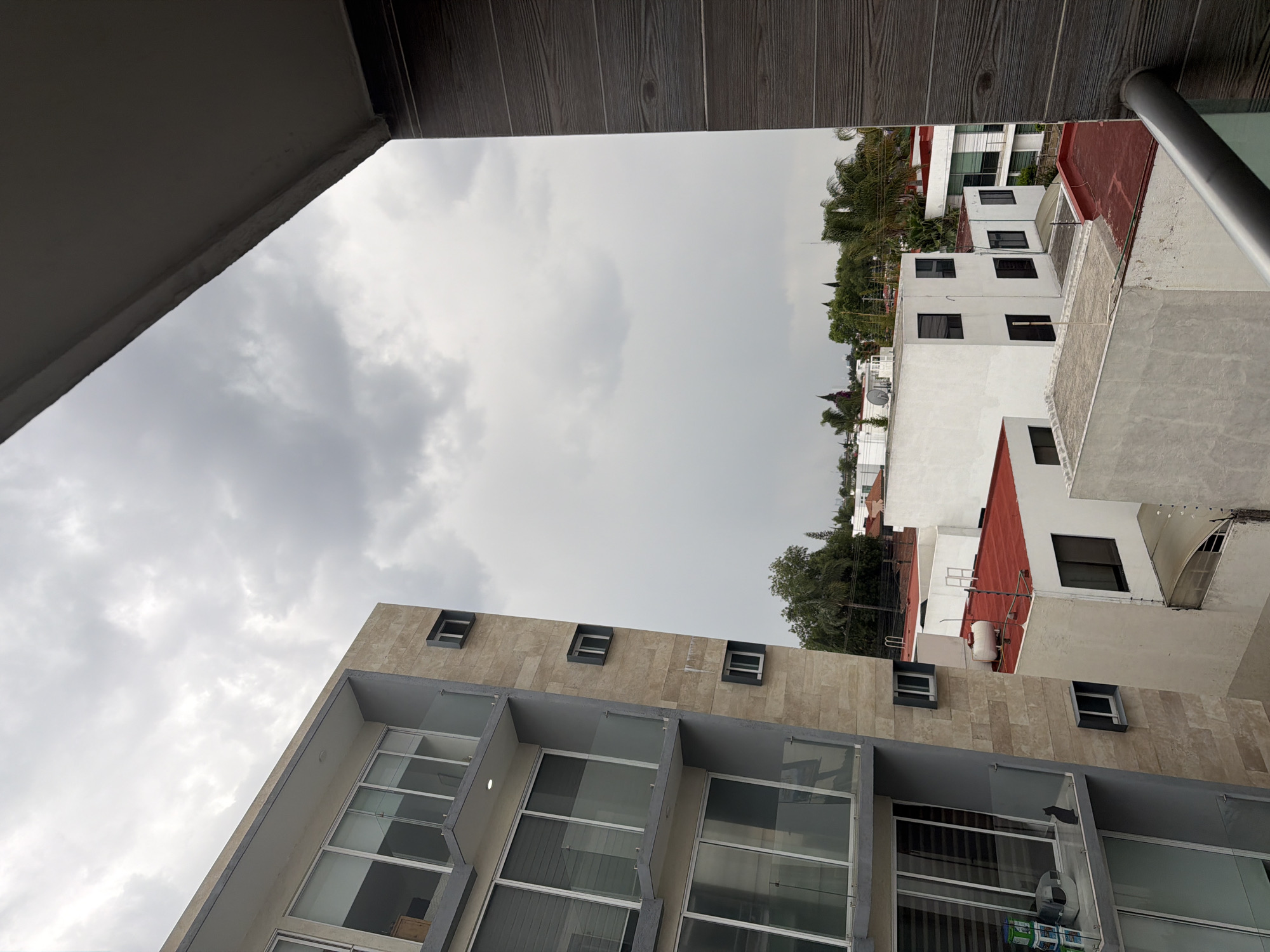 A low-angle, outdoor shot shows multiple buildings under a cloudy sky in San Andrés Cholula, Mexico. The foreground building has a tan, stone-like facade with large windows and a gray frame. Above this building are several white buildings with red roofs and black windows. Green trees are interspersed between the buildings. The sky is overcast with gray clouds. The image is framed by parts of the balcony above the vantage point.