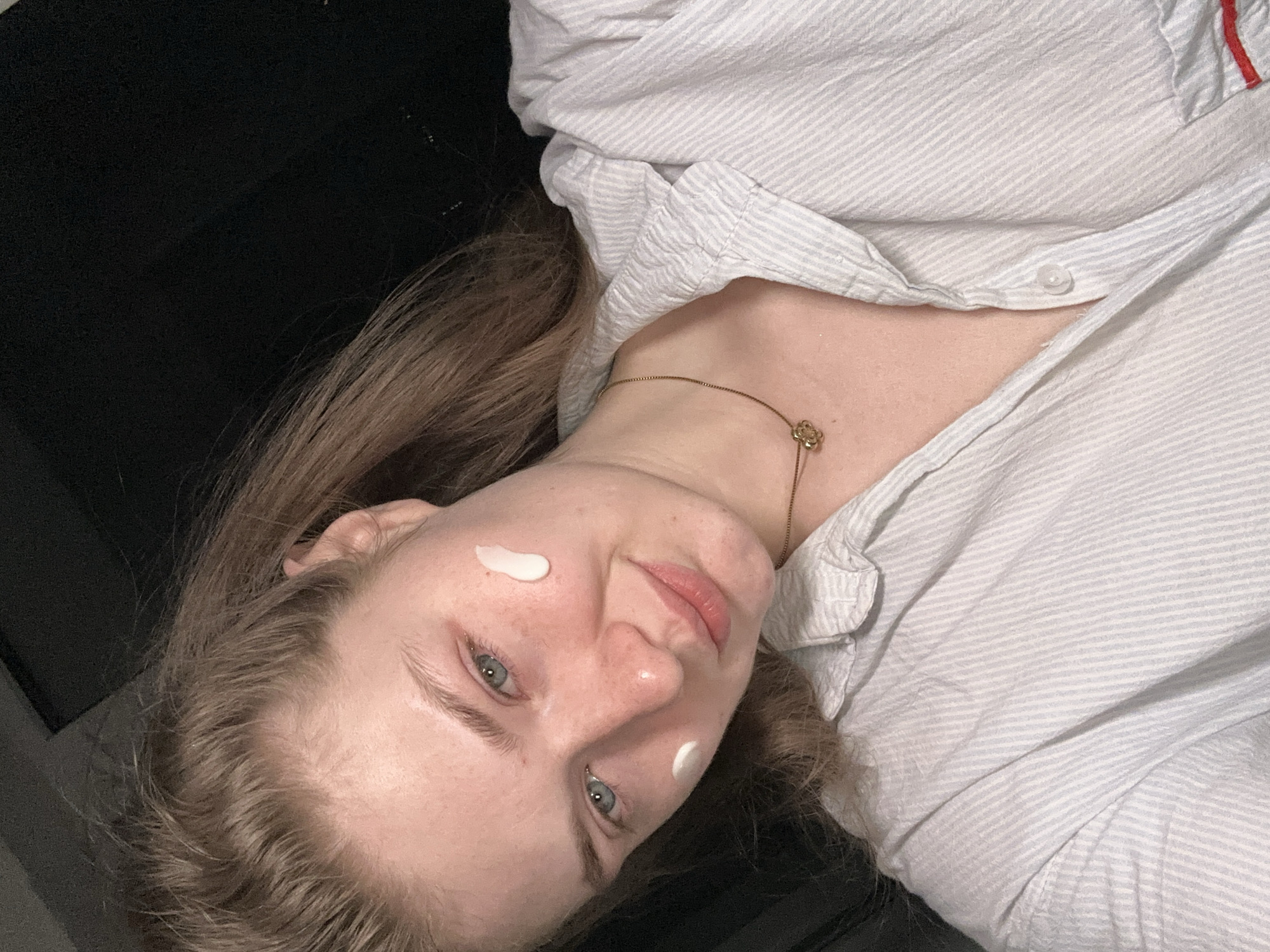 A fair-skinned young woman with light brown hair and blue eyes is pictured at an angle. She is wearing a striped button-down shirt and a delicate gold necklace with a floral pendant. She has two small dabs of white cream or lotion on her face, one on her cheek and one near her hairline. The background appears dark and out of focus. The location is not discernible from the image and there is no evidence of Kazan, Russia.