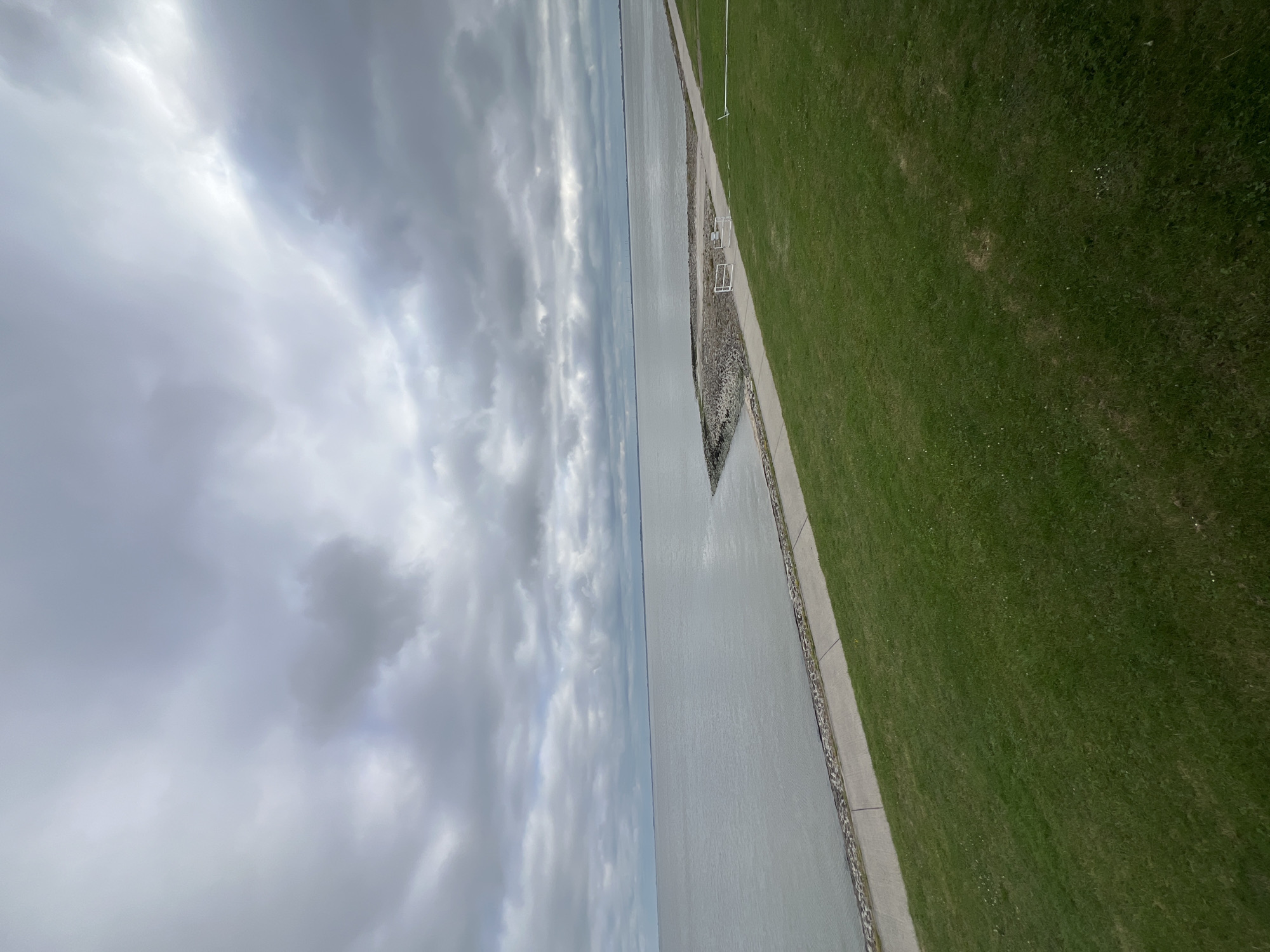 The image depicts a vast, calm body of water, likely the Jade Bay, with a cloudy sky overhead. The water meets a paved walkway, which is bordered by a well-maintained grassy area. The walkway has two white benches, suggesting a place for rest or observation. The scene is oriented such that the water occupies the left side of the frame and the grassy area the right, with the walkway acting as a boundary.
The sky is filled with a mix of dark and light gray clouds, indicating overcast or possibly impending rain. The overall mood is somewhat somber, fitting for a cloudy day. The light suggests it could be daytime, though the clouds obscure direct sunlight. There are no people visible in the image, giving a sense of solitude.
The setting appears to be a coastal or waterfront area, characteristic of Wilhelmshaven, Germany, a city known for its port and proximity to the sea. The presence of the walkway and benches suggests a public space designed for recreational use, possibly a promenade or a dike. The detailed texture of the grass and the stonework along the walkway are discernible. No visible text is present in the image.