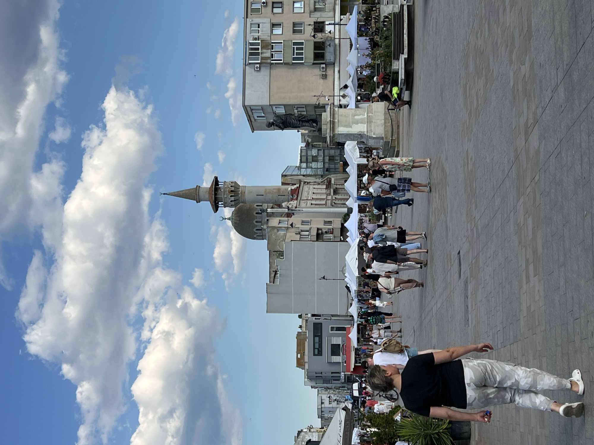 In Constanța, Romania, on a sunny day with scattered clouds, a bustling public square is captured. The foreground is dominated by a woman walking away from the camera, wearing a black t-shirt and camouflage pants. The middle ground reveals a lively scene with numerous people browsing stalls set up under white tents. A statue stands prominently on a pedestal, and behind it, a mosque with a tall minaret and a dome is visible, characteristic of the city's architectural blend. Buildings in various architectural styles, some modern apartment blocks and others older structures, form the backdrop. Street lamps and planters add to the urban setting. The ground is paved with stone tiles.