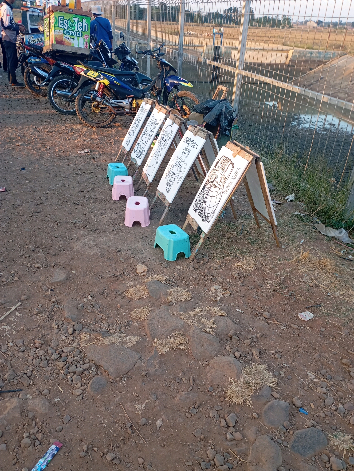 The image depicts an outdoor scene in Kecamatan Haurgeulis, Indonesia, likely a roadside or open area. In the foreground, several easels are set up with white canvases featuring black and white drawings. Some of the drawings appear to be of cartoon characters and vehicles. Three small, colored plastic stools (pink and turquoise) are placed in front of the easels, suggesting an area for people to sit and draw or color. The ground is covered with dirt, gravel, and sparse dry grass.
Behind the easels, several motorcycles are parked in a row. The motorcycles are various colors, including blue, black, and yellow accents. One of the motorcycles has the number "23" on its side. To the far left, a cart is visible, with a sign on it that says "Es Teh POCI". A person wearing a white hat and blue shirt stands near the cart. Another person, dressed in dark clothing, is also present near the cart, looking towards it.
In the background, a metal fence runs across the scene. Beyond the fence, a body of water, possibly a canal or irrigation ditch, is visible, and further back, a dry, cultivated field stretches into the distance. The sky suggests it is either early morning or late afternoon, with warm lighting. The overall atmosphere suggests a casual, possibly recreational setting, perhaps a temporary stall set up for selling drinks and offering drawing activities.