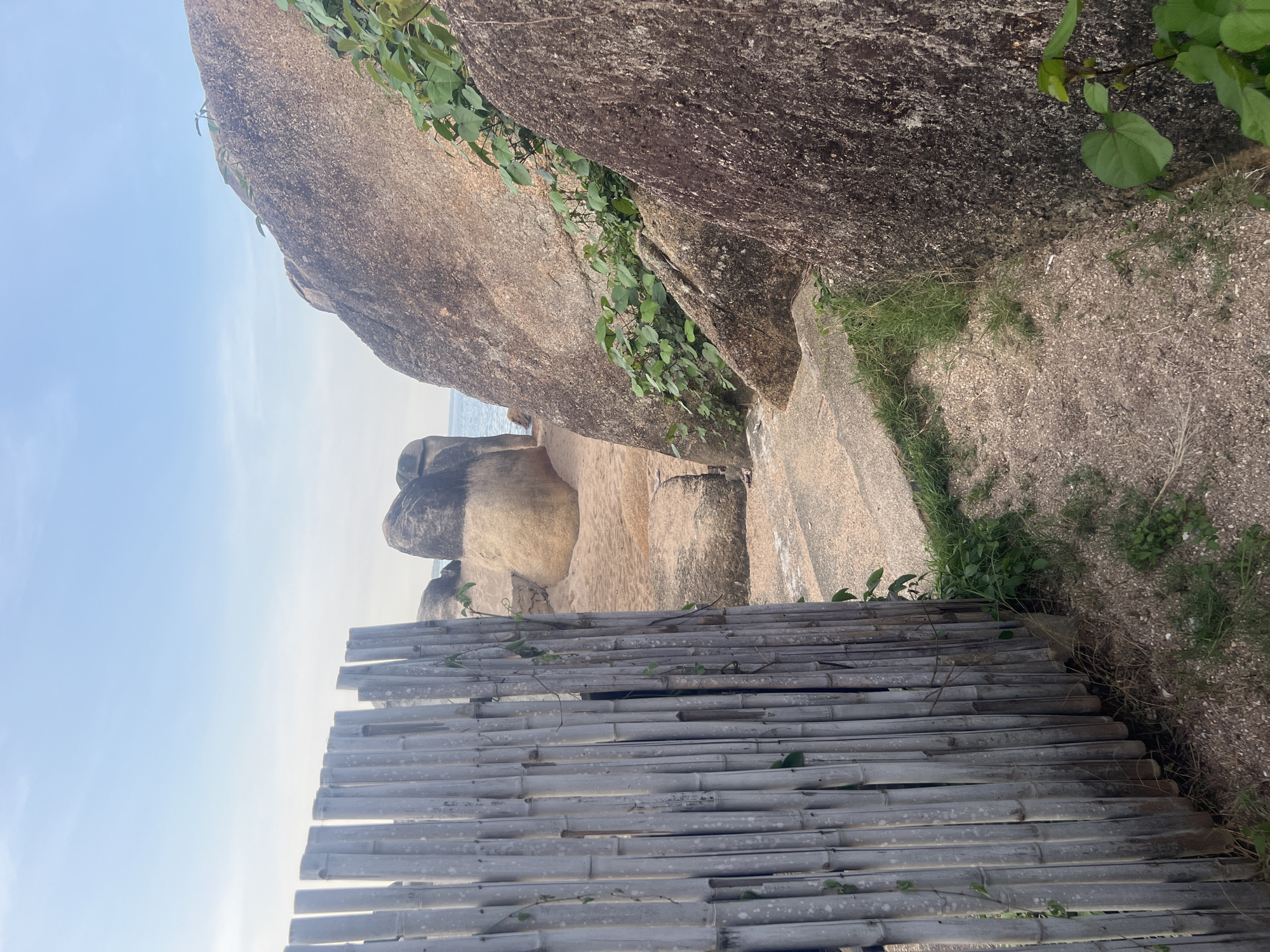The image shows a path leading through a rocky area on the coast of Ko Samui District, Thailand. On the left, a weathered bamboo fence partially obstructs the view, its vertical slats weathered and grey, with a few green vines clinging to it. The path itself is made of sand and small stones, with patches of green grass growing alongside. To the right of the path, large, smooth boulders dominate the scene. The largest boulder is in the foreground, its surface rough and dark. Further back, more rounded, lighter-colored boulders are visible, with vegetation growing between them. In the background, the calm sea meets a hazy sky. The scene is tranquil and natural, suggesting a secluded spot on the island. There are no people or animals visible in the image.