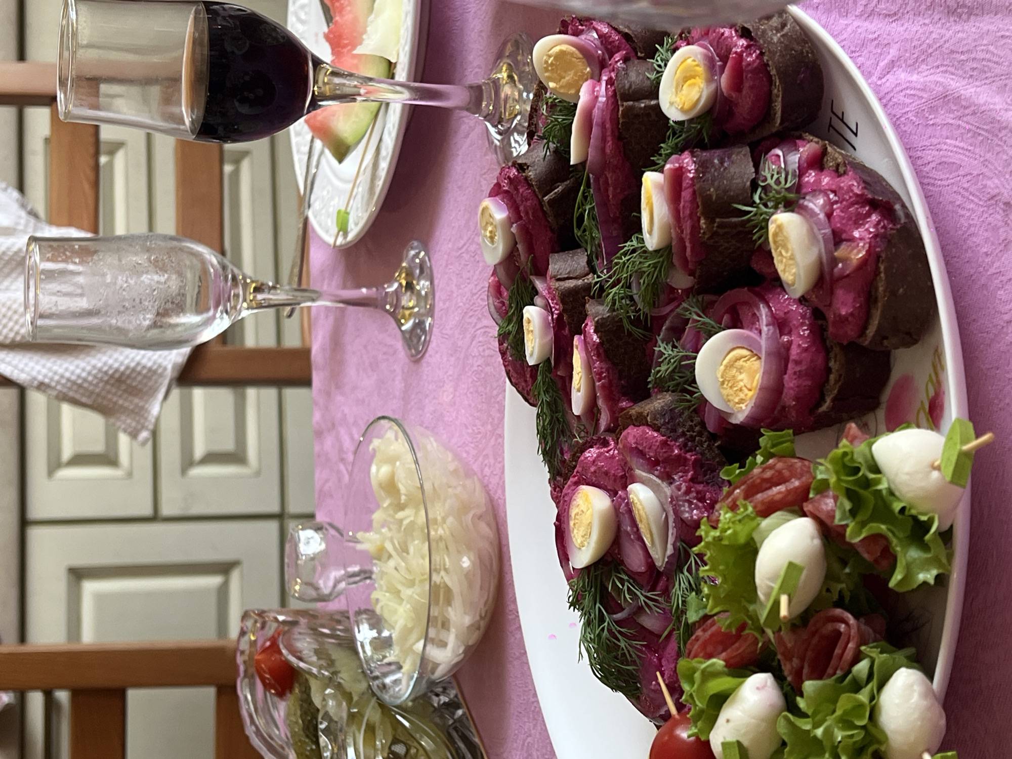 This image depicts a table set with a meal, likely in a home dining area within Mezhdurechenskiy, Russia. A pink tablecloth covers the table, on which several dishes are arranged. In the foreground, a white platter holds numerous open-faced sandwiches topped with a vibrant pink spread, likely beet-based, along with sliced hard-boiled quail eggs, red onion rings, and fresh dill. To the left of the platter, skewers with cherry tomatoes, small white mozzarella balls, and rolled salami slices are presented. Behind these dishes, a clear glass bowl filled with shredded cabbage salad is visible, and further back, a crystal bowl contains pickles and a tomato. Two stemmed glasses are present: one is empty and appears slightly frosted, while the other contains red wine. A white plate with slices of watermelon sits between the glasses. The background reveals a wooden chair and kitchen cabinetry, suggesting an indoor domestic setting. There are no people visible in the image.