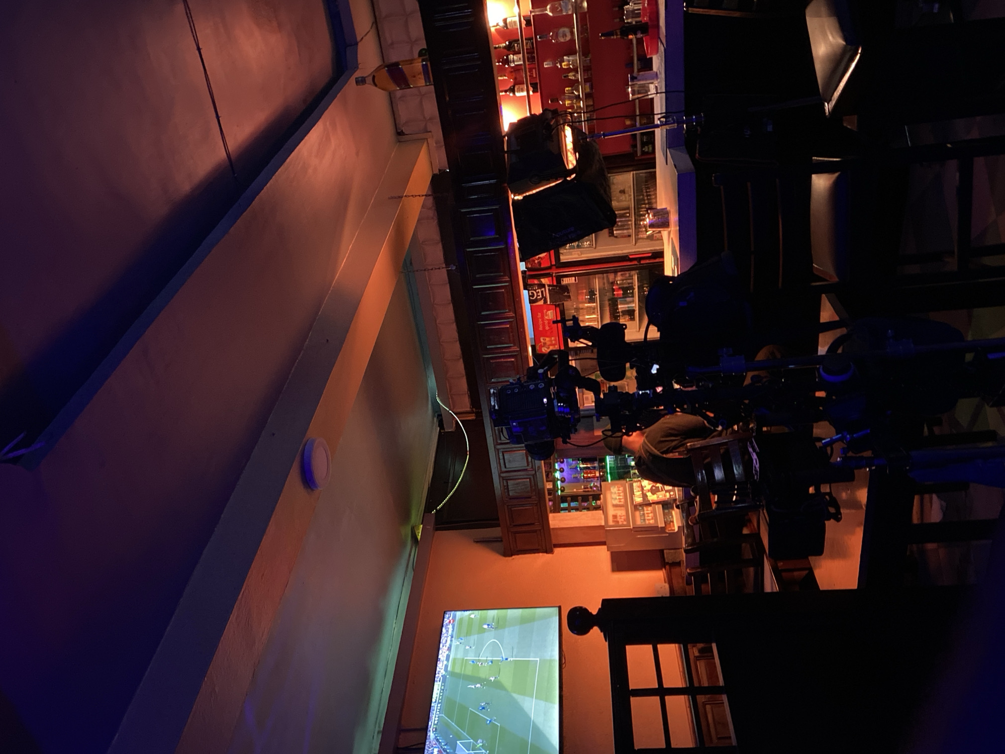 The image depicts a scene inside a bar, possibly in Lagos, Nigeria. A football match is being televised on a large screen mounted on the wall. A camera crew is filming the patrons watching the game. A bar counter, with bottles and glasses is visible behind them. The lighting is subdued, creating a relaxed atmosphere. Dark chairs are in the foreground, with people seated and watching TV.