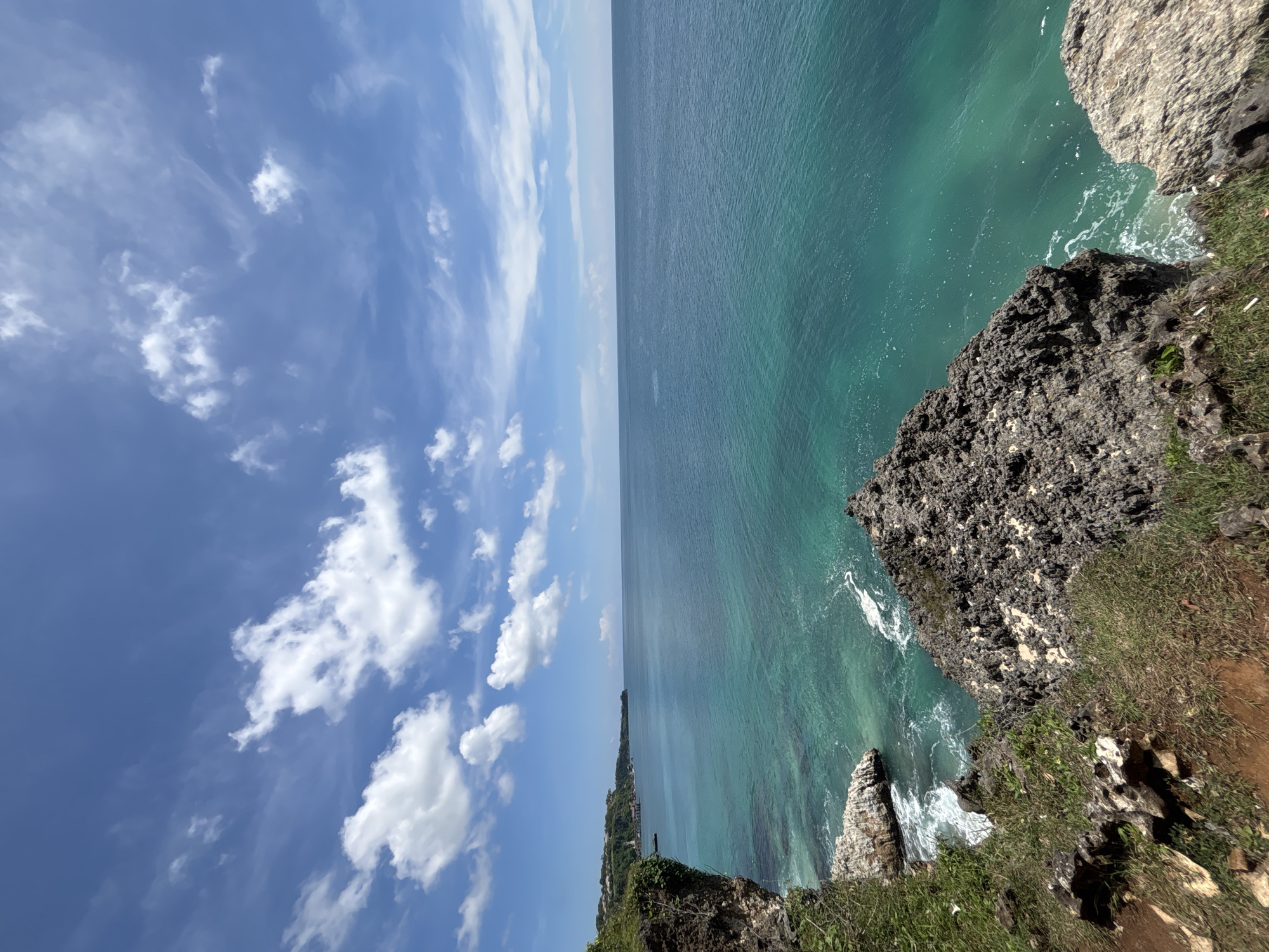 The image shows a coastline in Unknown, Indonesia. The scene is divided roughly in half, with the left side showing a bright blue sky with scattered white clouds, and the right side showing the ocean and rocky cliffs. The ocean is a vibrant turquoise color, with visible wave action near the cliffs. The cliffs are composed of dark, textured rock with patches of green vegetation. The image appears to have been taken from a high vantage point on the cliffs, looking out over the ocean.