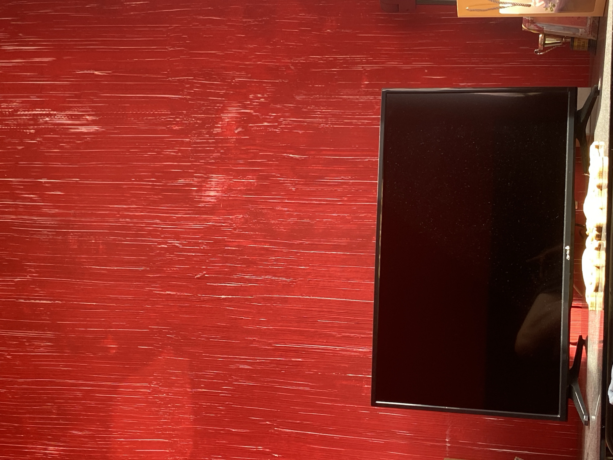 The image displays an indoor scene, likely a room in Troyan, Bulgaria, characterized by a strikingly red wall that occupies most of the left and central portion of the frame. This wall features a textured, distressed finish with numerous fine, horizontal, lighter red and white brushstroke-like lines, giving it a raw or artistic appearance.
To the right, a black, rectangular flat-screen television is prominently positioned. Its screen is dark, reflective, and appears to have a fine layer of dust or smudges. A faint white logo, possibly "LG," is discernible on the bottom right corner of the screen. The TV is supported by a black stand, partially visible beneath its right side.
Above the television and extending towards the top right corner of the image, parts of a shelf or surface are visible. On this surface, a small, golden trophy with a dark base can be seen, its details unreadable. Next to the trophy, a segment of a gift bag or box, featuring a floral pattern and a string handle, is also present.
The scene is static, with no activity taking place. The lighting suggests an indoor setting, but there are no specific cues to indicate the time of day or external weather conditions. The overall impression is that of a decorated interior space with a bold color scheme.