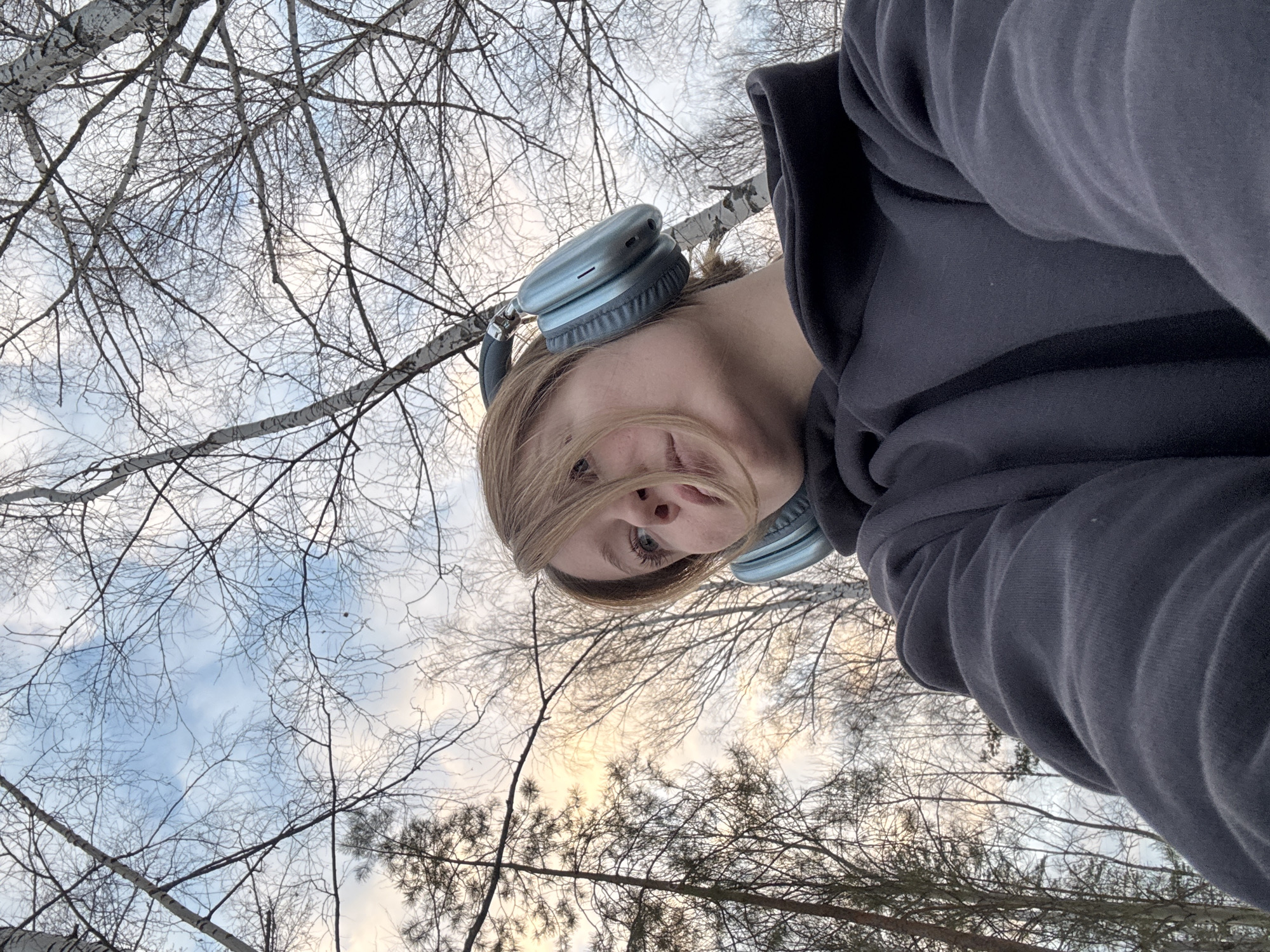 An individual is depicted from a low-angle perspective within a wooded environment in the city of Chaikovsky, Russia. The subject, appearing to be a young adult female with light brown hair, is wearing a dark gray hooded sweatshirt and light blue/silver over-ear headphones. Her head is tilted slightly upwards and to her left, with her gaze directed in the same general direction. The background features a dense collection of trees, primarily bare deciduous trees with visible white bark, likely birches, and several dark green coniferous trees with needles, particularly on the left side of the frame. The sky above is partially cloudy, showing patches of light blue interspersed with muted white, yellow, and orange hues, indicative of early morning or late afternoon light conditions.