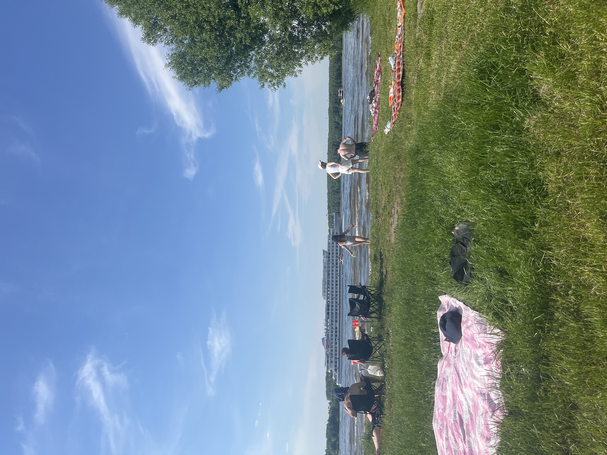 The image captures a sunny summer day at a riverbank in Kapustino, Russia, featuring a wide river and a grassy shore. The sky is clear blue with sparse, wispy white clouds.
In the mid-ground, a large, multi-deck white passenger ship with red accents on its smokestack travels left to right across the river. Several smaller boats are visible further in the distance.
On the left side of the foreground, a pink and white patterned blanket is spread on the green grass, with a dark blue object resting on it. To the right of the blanket, another dark object lies on the grass. Closer to the water's edge, one person lies on their stomach on the grass. Next to them, two individuals are seated in black folding chairs, facing the river; one wears a dark blue head covering. Between these chairs, a small portable table holds red cups. A third empty black folding chair is positioned further to the right. A child in a light-colored swimsuit stands in the shallow water, facing the passing ship.
On the right side of the foreground, an orange, red, and white patterned blanket is spread on the grass, covered with various items of clothing or towels. Two adults stand at the water's edge, their backs partially to the viewer, observing the water. One wears white shorts, a white top, and a white baseball cap; the other wears dark shorts. A large tree with dense green foliage occupies the far right of the frame. The opposite bank of the river is lined with a dense treeline.