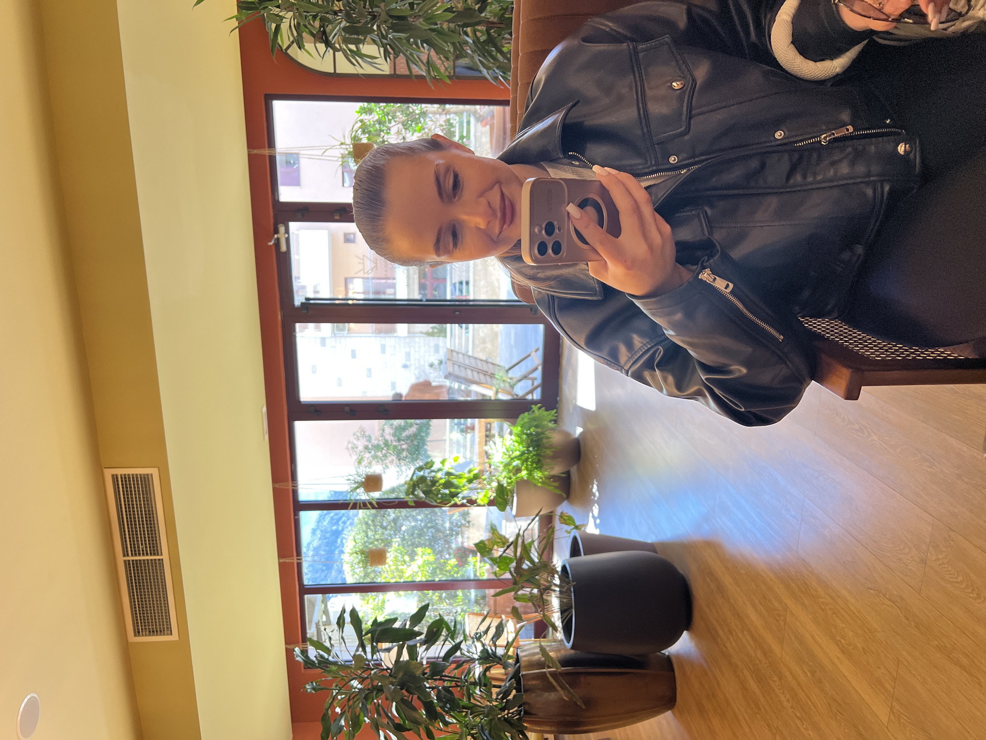 A woman in a black leather jacket sits indoors, holding a smartphone to take a selfie. Large windows behind her reveal a view of Boreti, Montenegro, with plants placed in front of the windows, softening the urban scene. The setting appears to be a cafe or restaurant interior.